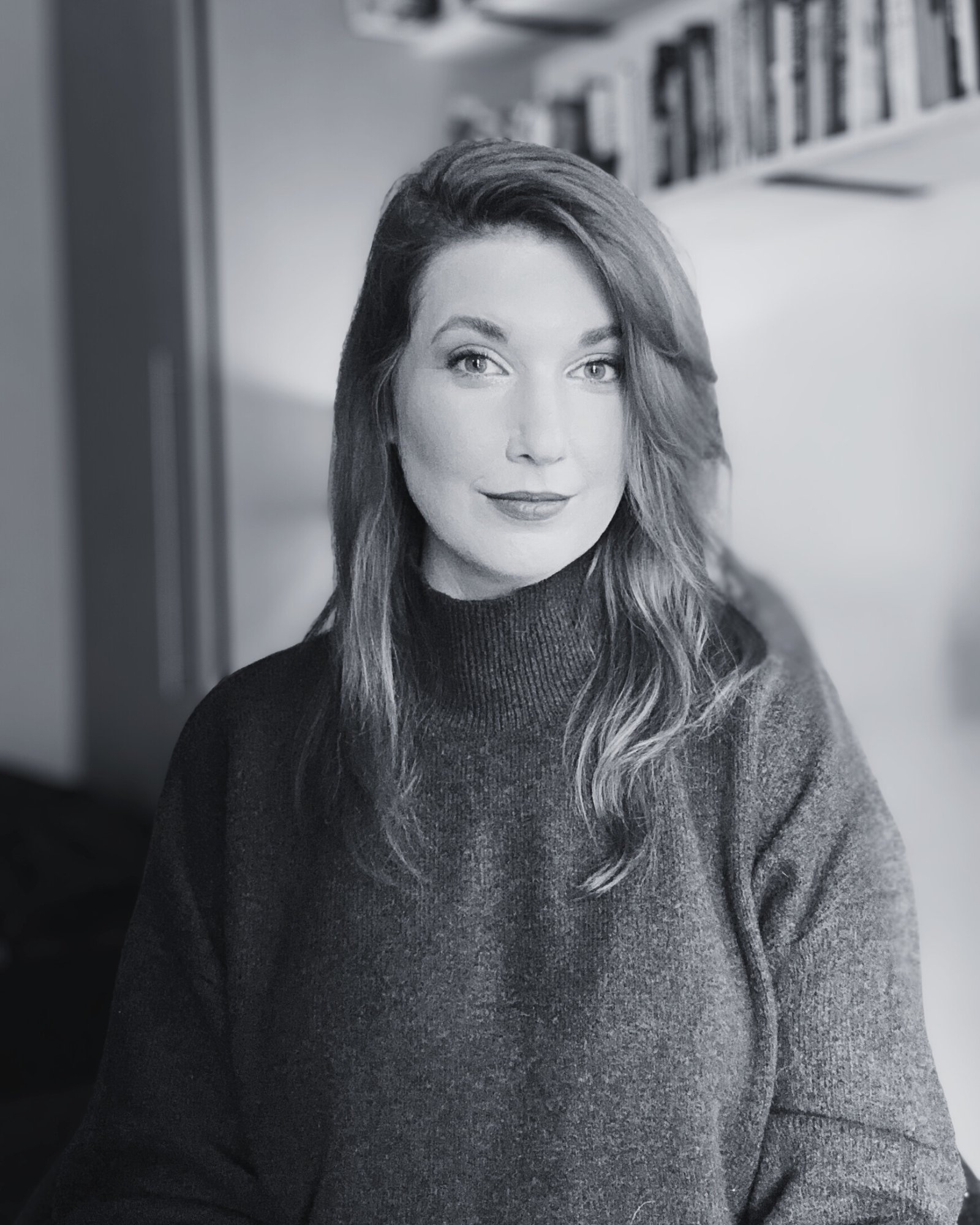A woman with long hair and a turtleneck sweater sitting indoors, looking at the camera with a slight smile. The background features a bookshelf.