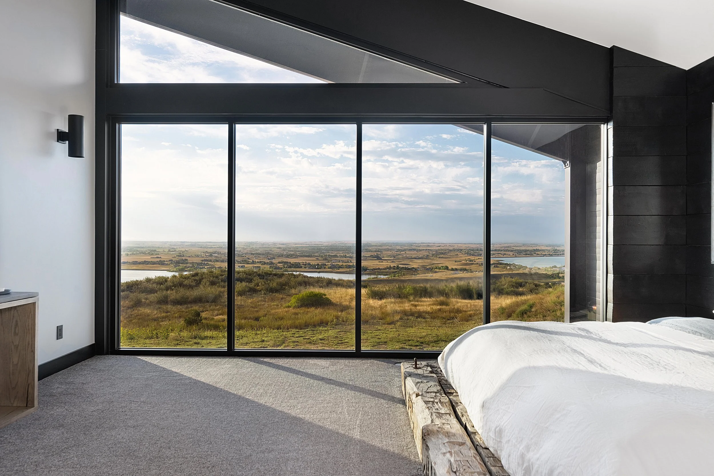 Modern bedroom with large glass windows showcasing a scenic landscape view, featuring open fields, a river, and a cloudy blue sky.