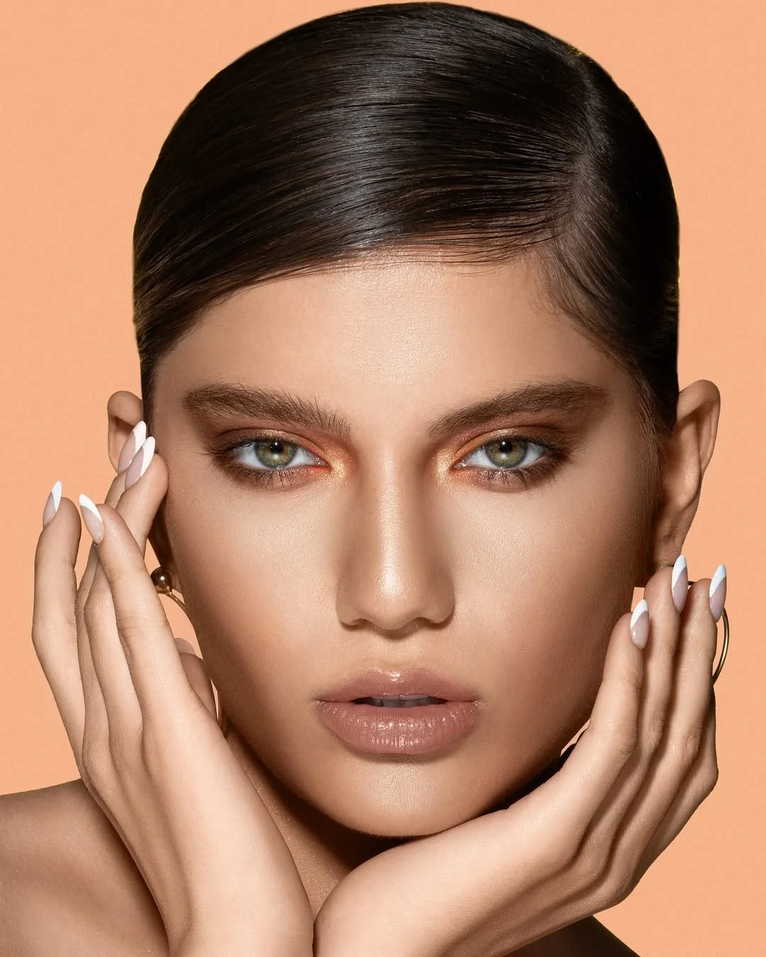 Close-up portrait of a person with slicked-back hair and neutral makeup, featuring prominent eye shadow and glossy lips, holding hands near the face against a peach-colored background.