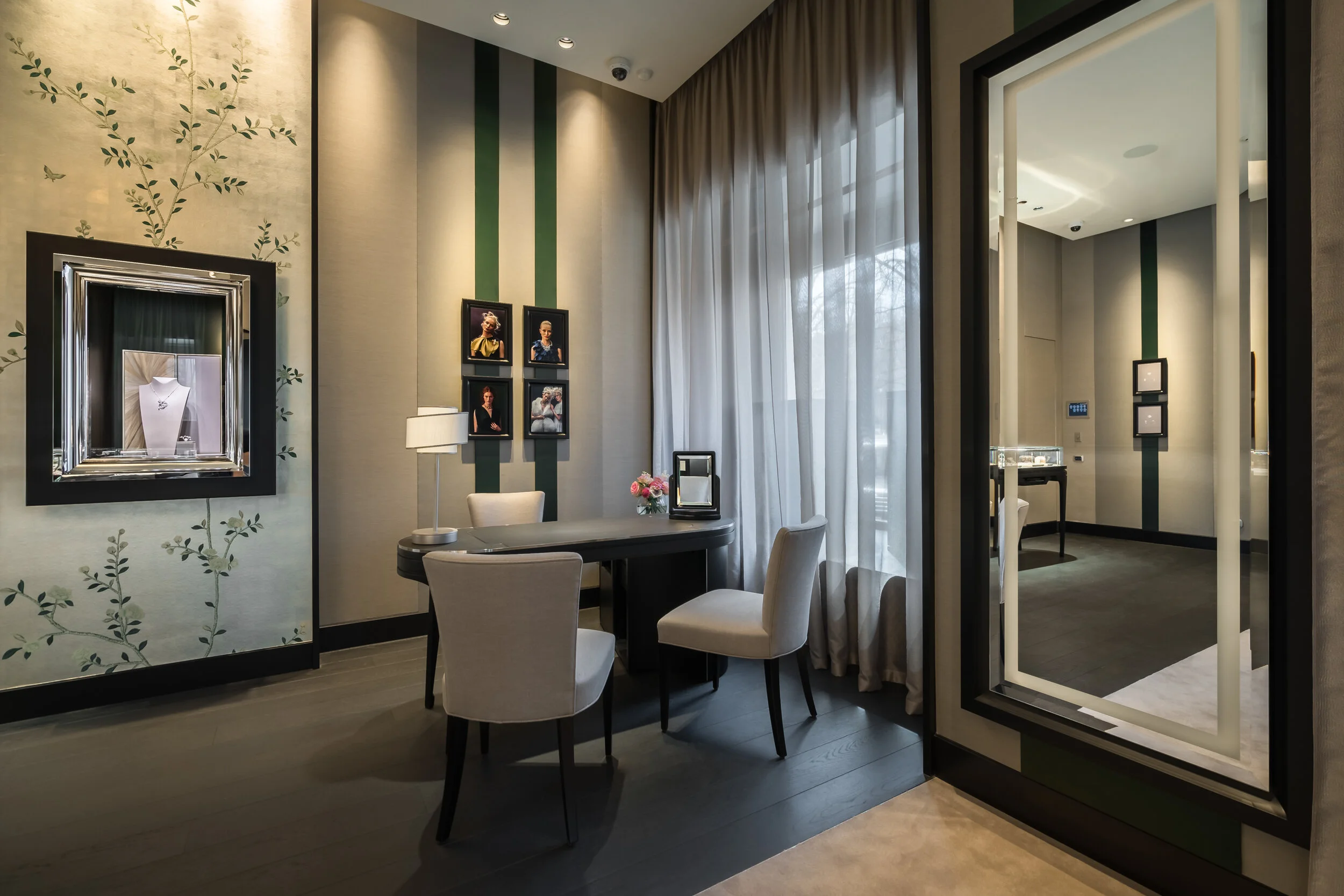 Elegant interior of a jewelry store consultation room with a table, chairs, floral wallpaper, framed pictures, and a window with sheer curtains. A jewelry display is visible through a doorway.