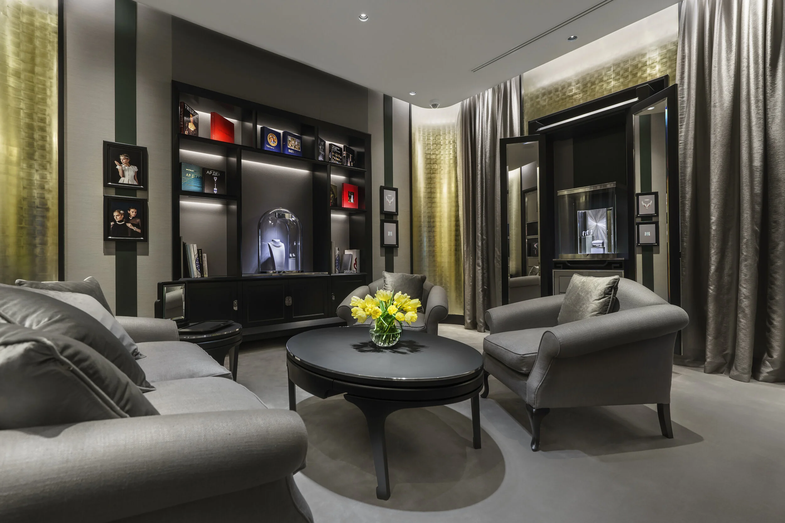 Luxurious living room with gray armchairs, a round table, and a vase of yellow tulips. Shelving with decorative items and framed photos on display. Soft lighting creates an elegant ambiance.