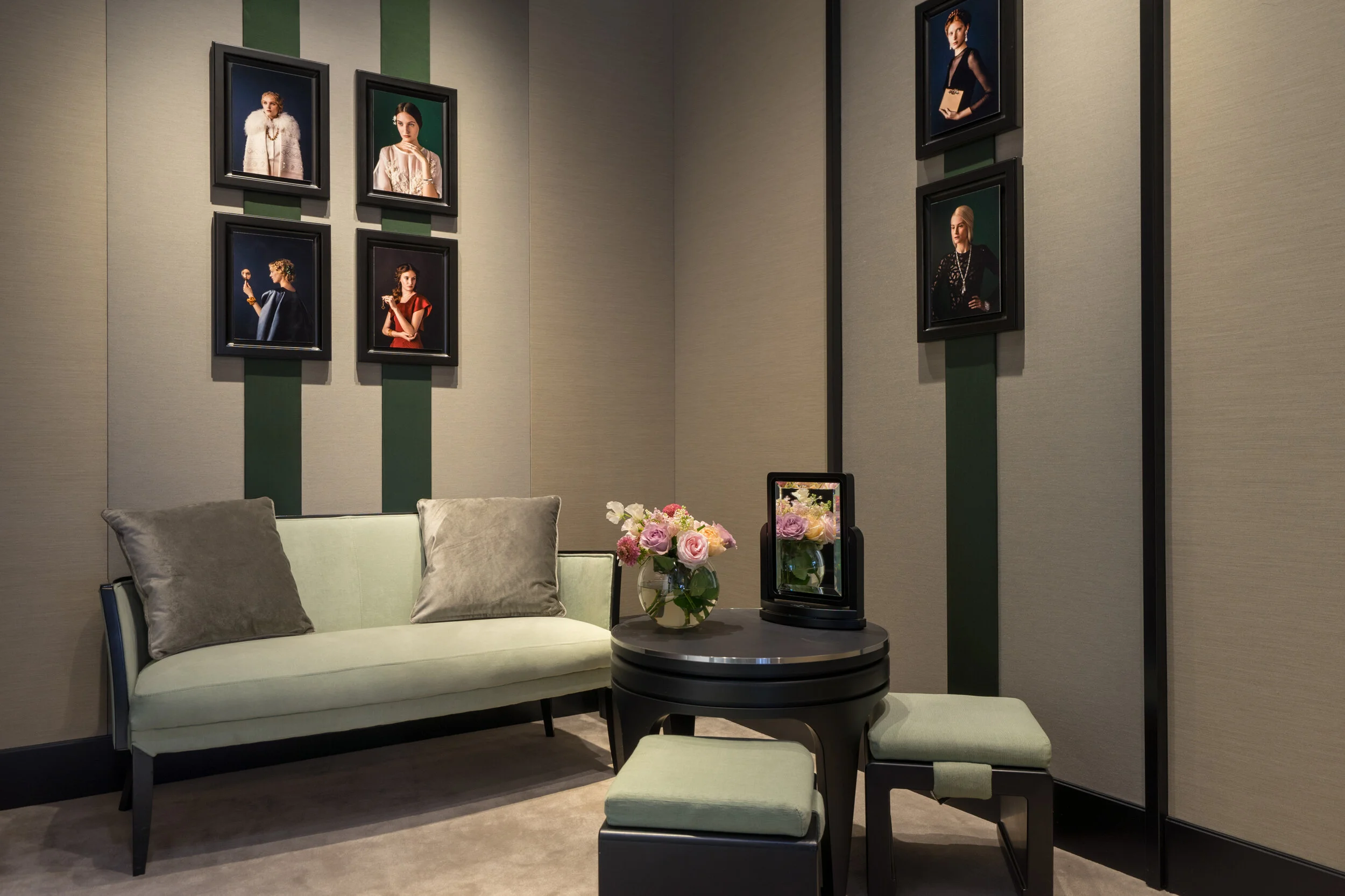 A stylish room with a light green sofa, two matching cushions, a round black table with a floral arrangement, and framed portraits on the walls.