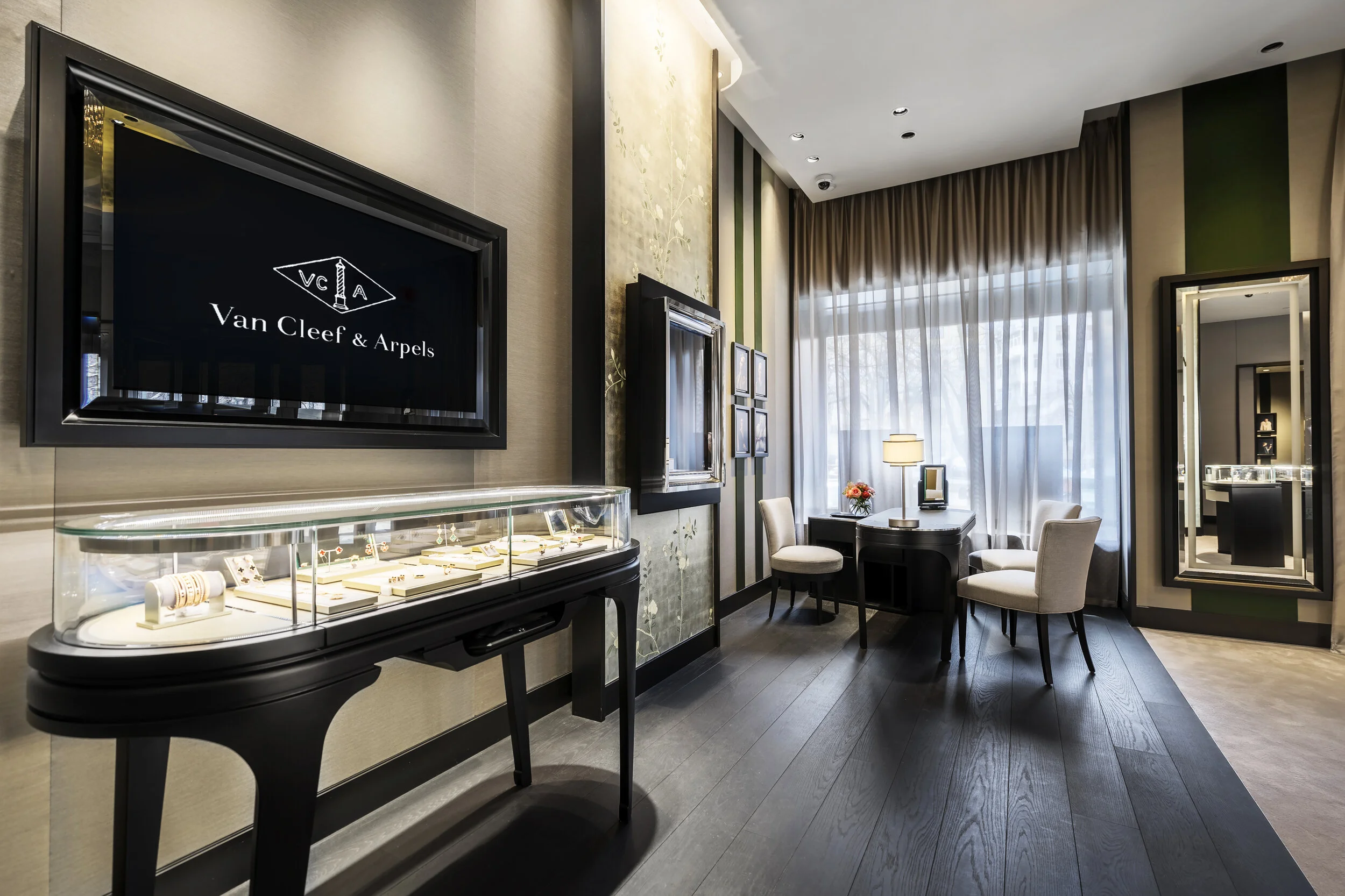 Interior of a luxury jewelry boutique featuring display cases with jewelry, elegant chairs, a desk with a lamp, and a Van Cleef & Arpels logo on the wall.
