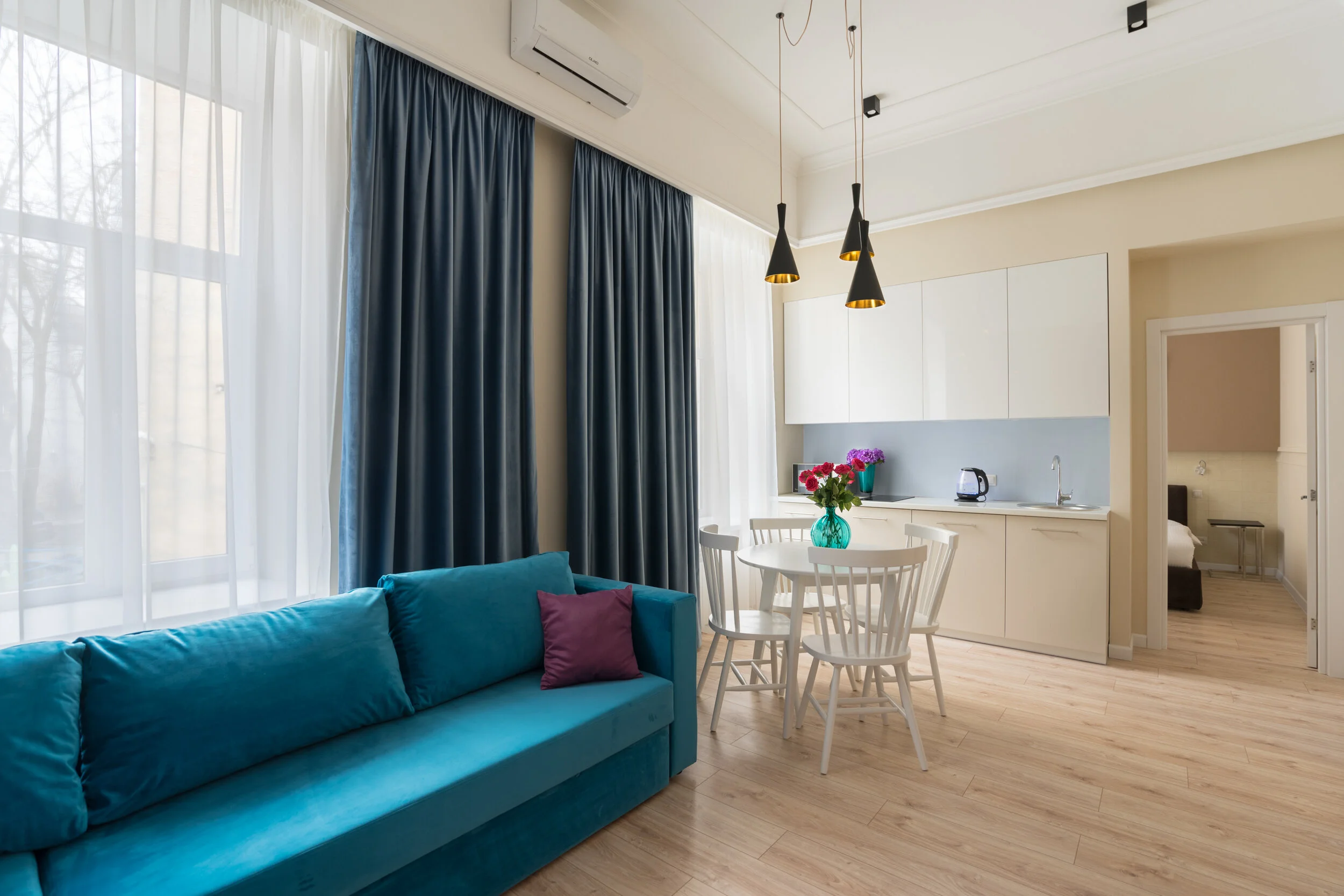 Modern living room and kitchen with blue sofa, white dining table, and chairs. Large windows with blue and white curtains. Hanging lights and a decorative vase with flowers on the table.