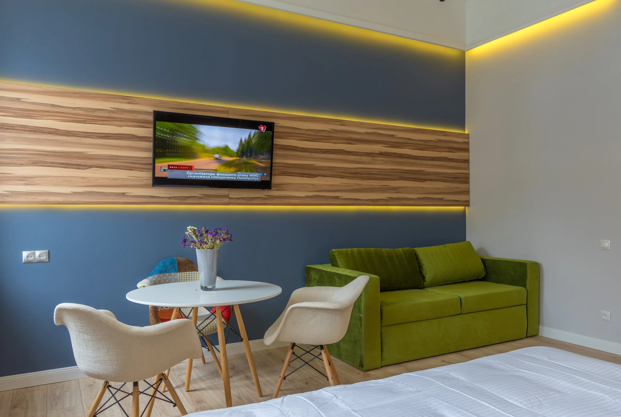 Modern living room with green sofa, white chairs, round table, wall-mounted TV, and wood accent paneling.