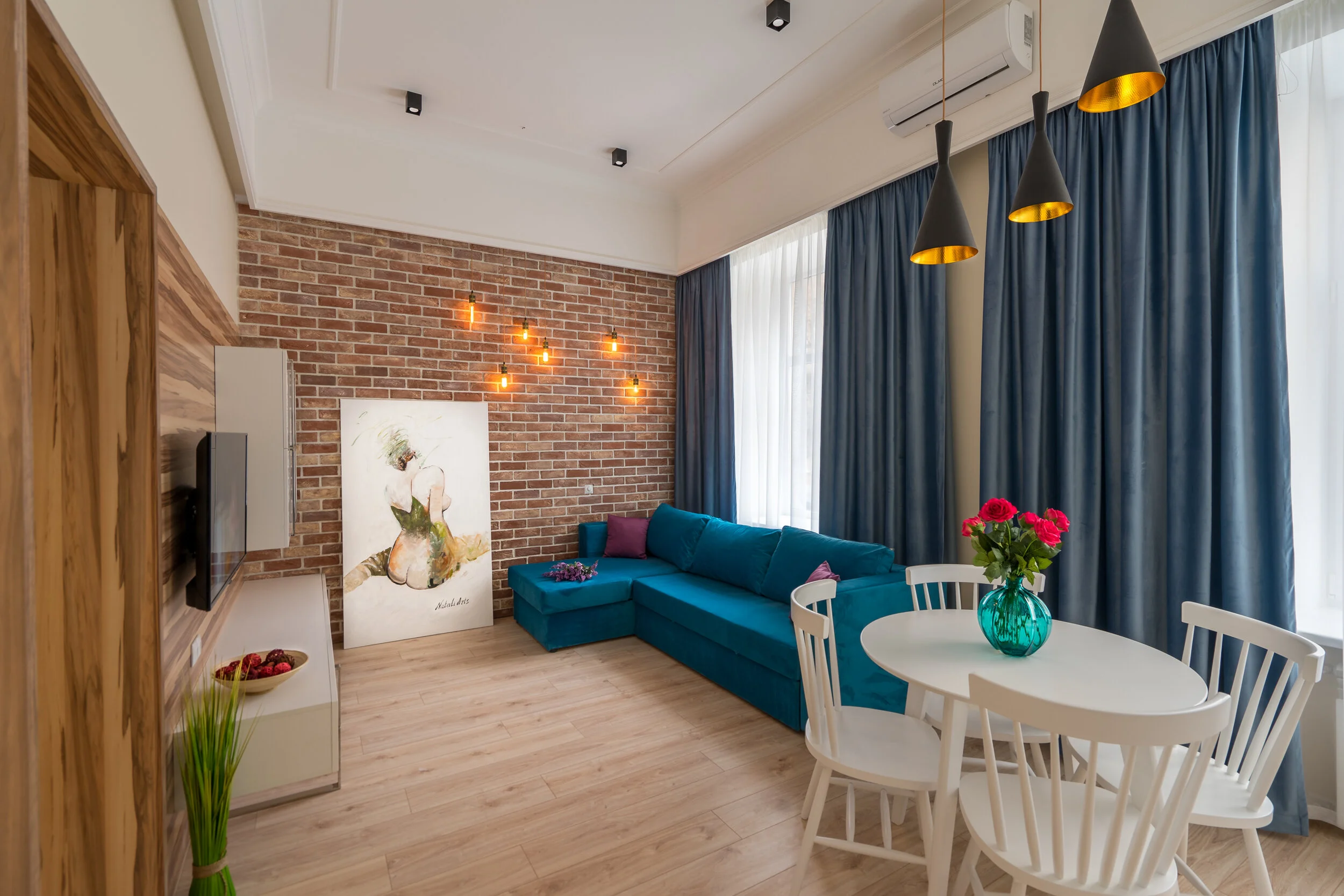 Modern living room with brick accent wall, blue sectional sofa, white round dining table with chairs, vibrant pink flowers in a teal vase, pendant lights, and large windows with blue curtains.
