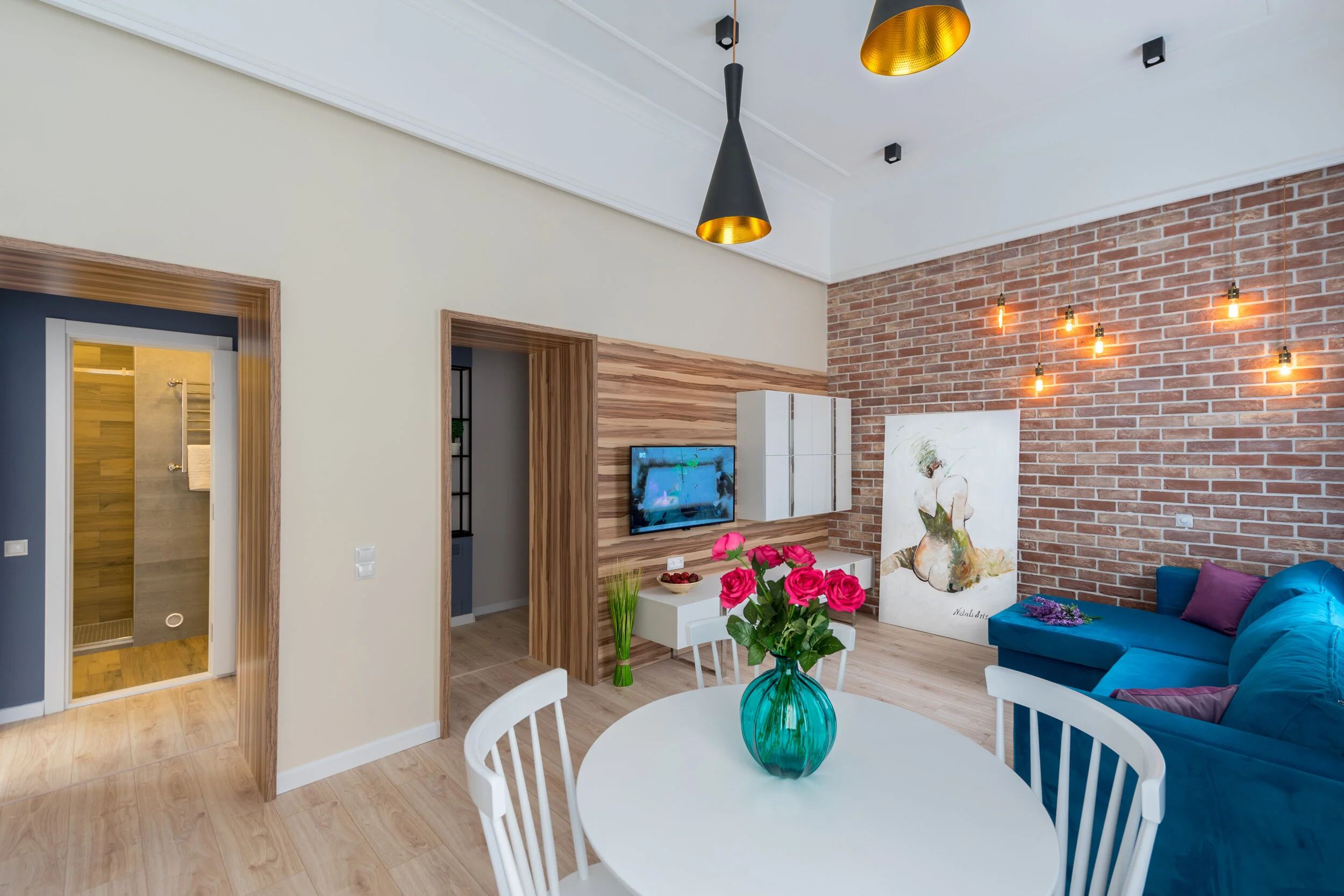 Modern living room and dining area with a blue sofa, brick accent wall, and a round white table with pink roses in a blue vase. A wall-mounted TV and decorative artwork enhance the aesthetic.