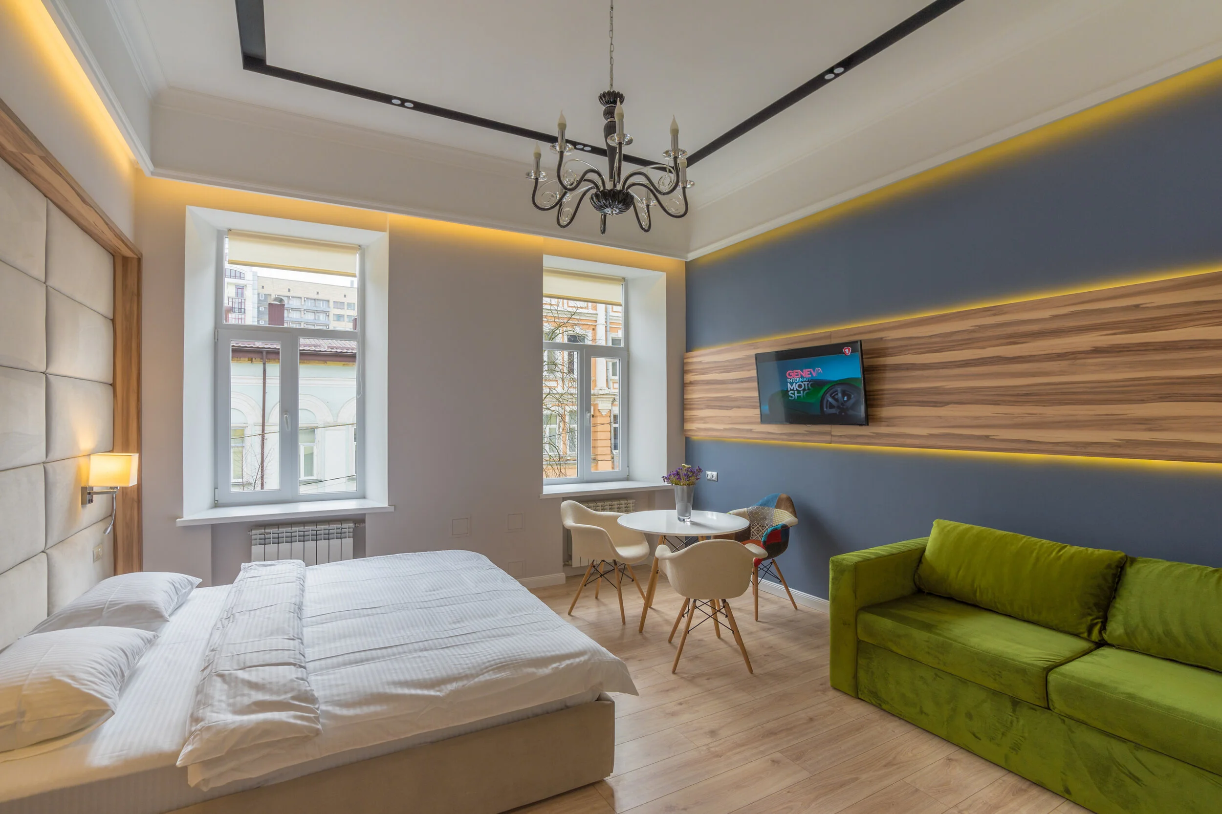 Modern studio apartment interior with a bed, green sofa, round dining table with chairs, wall-mounted TV, and decorative chandelier.