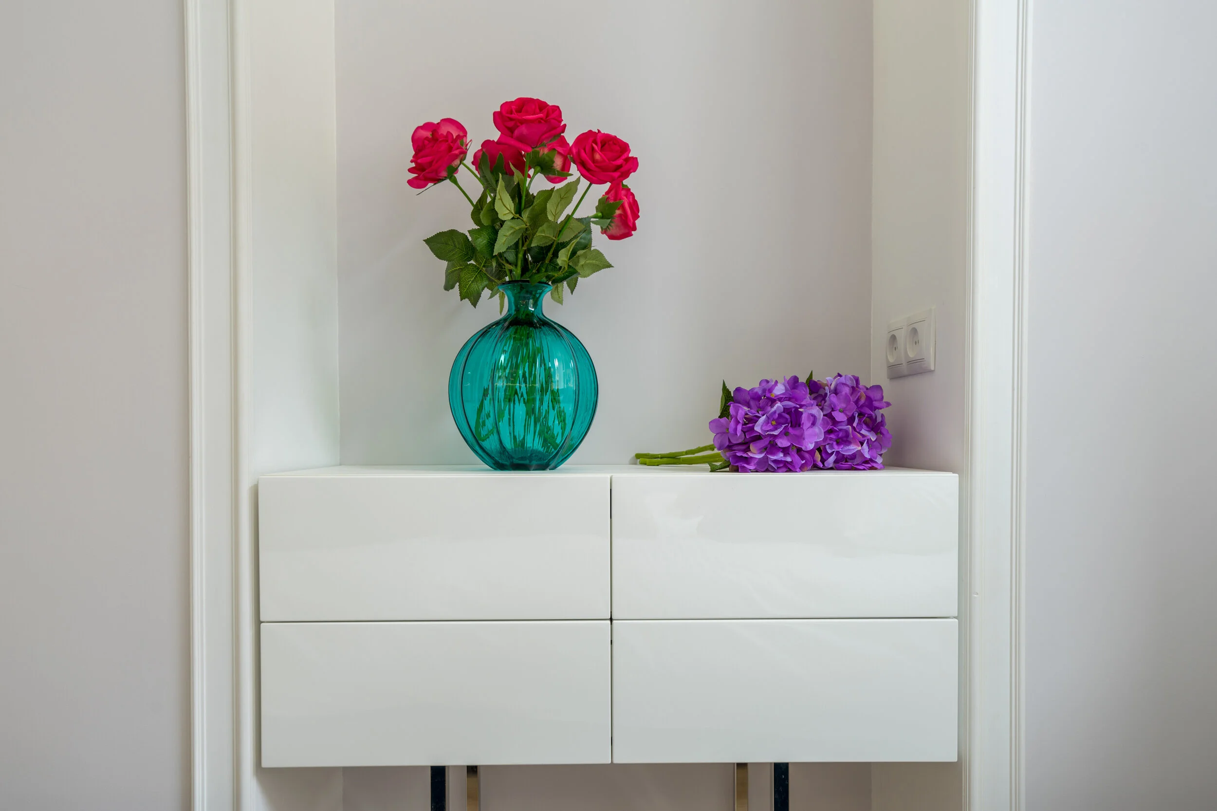 Pink roses in a blue glass vase on a white cabinet with purple flowers beside it.
