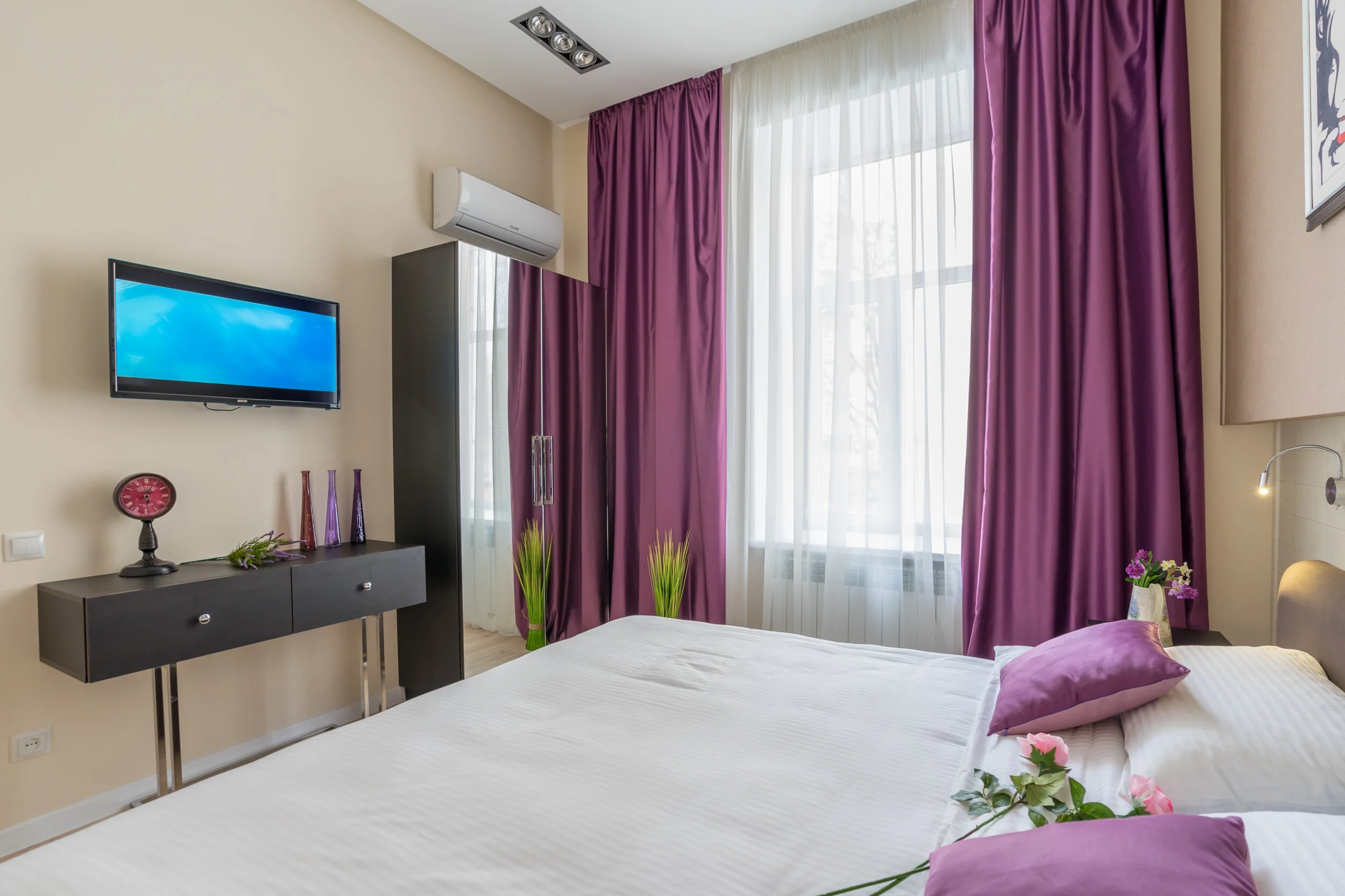 Modern bedroom with a double bed, purple curtains, TV, and wall-mounted air conditioner. The room includes a dresser with decorative items and flowers on the bed.