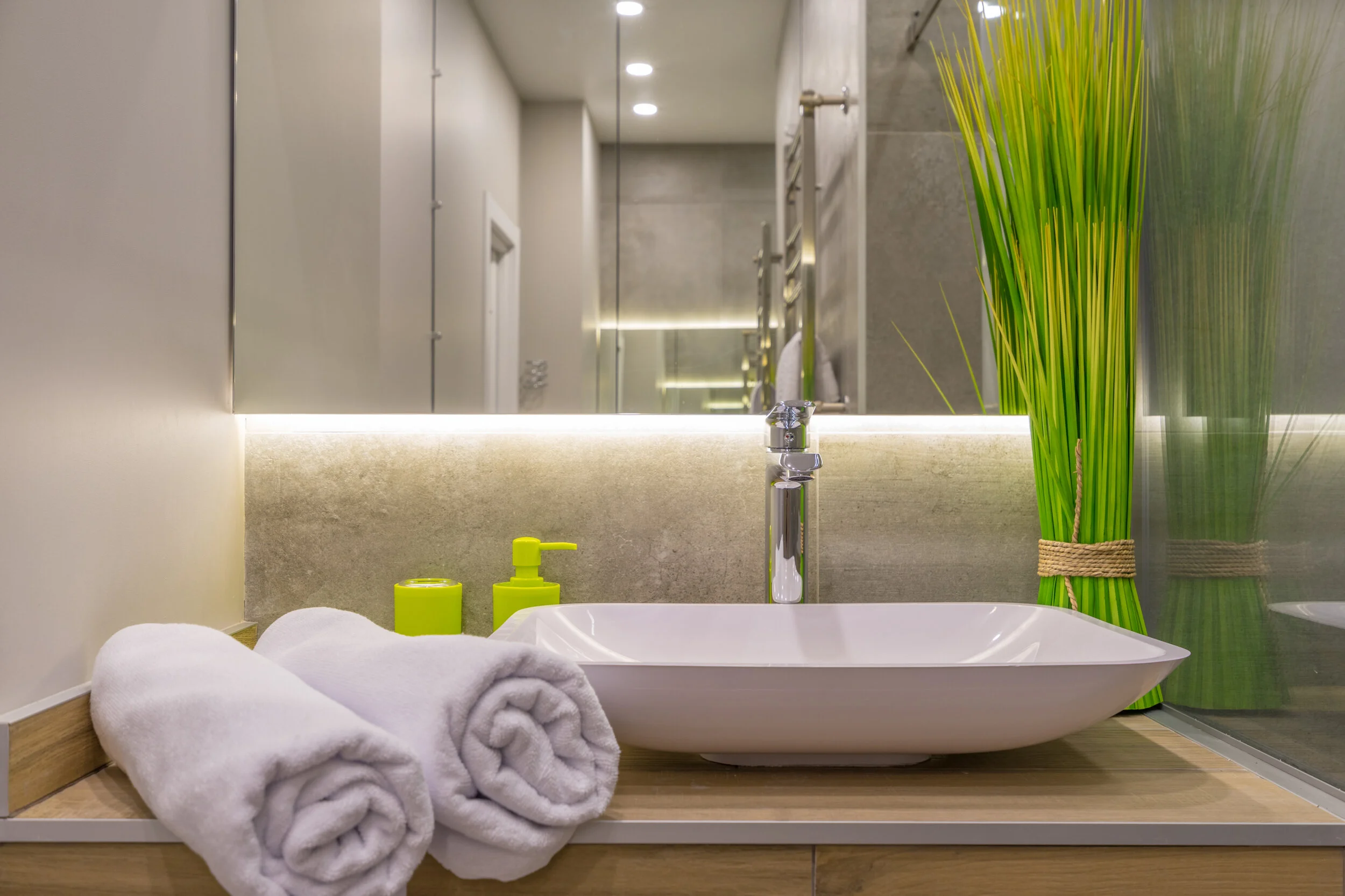 Modern bathroom with white square sink, chrome faucet, two rolled white towels, green soap dispenser, and tall decorative grass in a vase.
