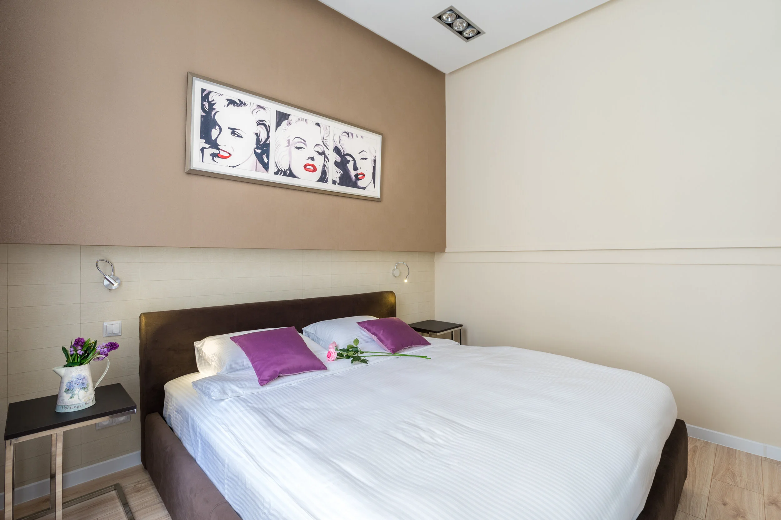 Modern bedroom with double bed, purple pillows, and artwork of a famous person's face above the bed. Side tables with a flower vase and wall lamps. Light wood flooring and neutral-colored walls.