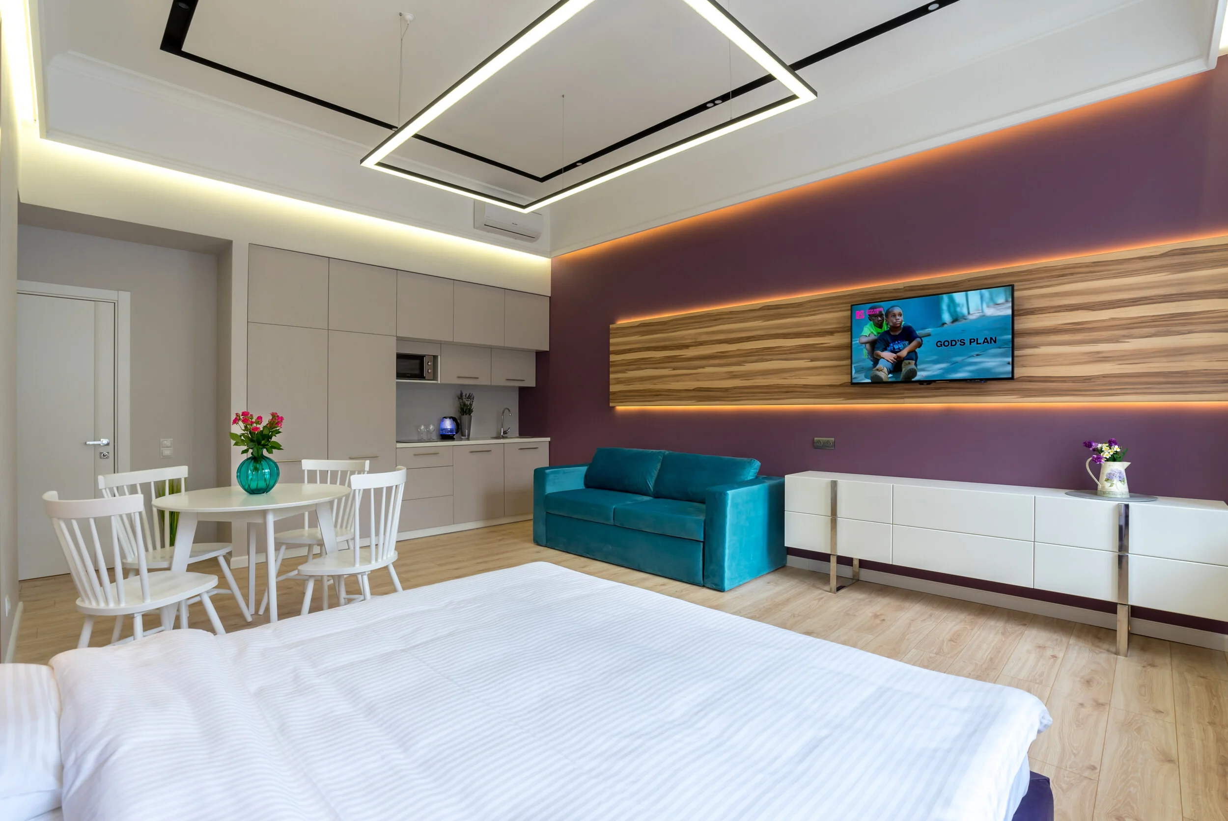 Modern studio apartment interior with kitchenette, dining area, and living space. Features white round table with chairs, teal sofa, and TV on a wooden accent wall. Minimalist design with purple and beige decor, wooden flooring, and ambient lighting.