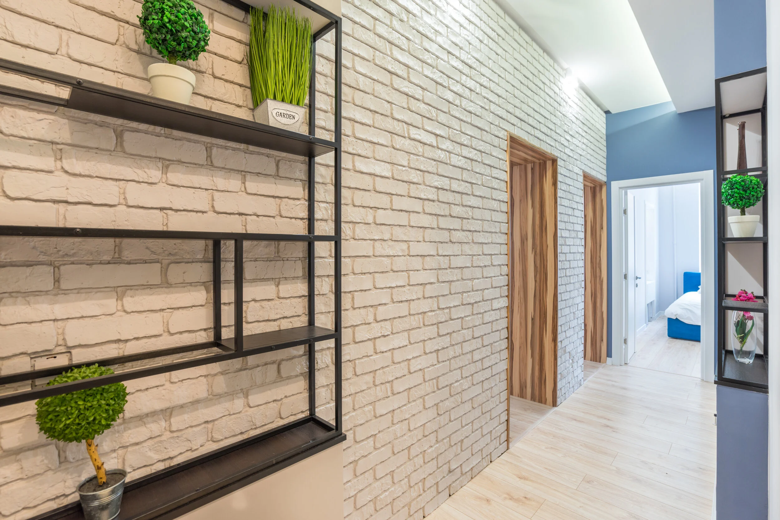 Modern hallway with white brick walls, wooden doors, and black metal shelves adorned with potted plants and decorative items.