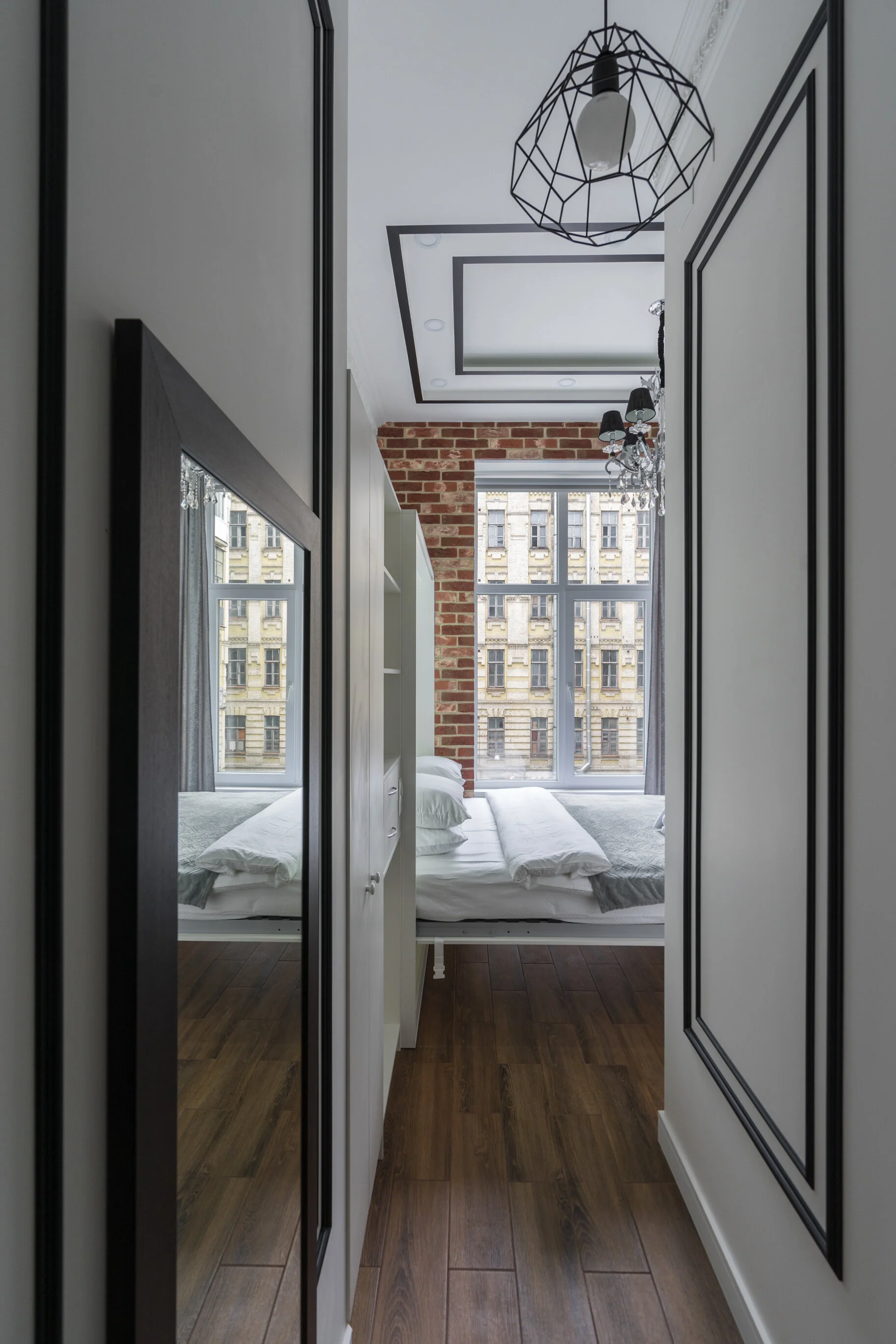 Small modern bedroom with brick accent wall, large window, and geometric pendant light. Includes a bed with white linens and two pillows, a mirrored wardrobe, and dark wood flooring.