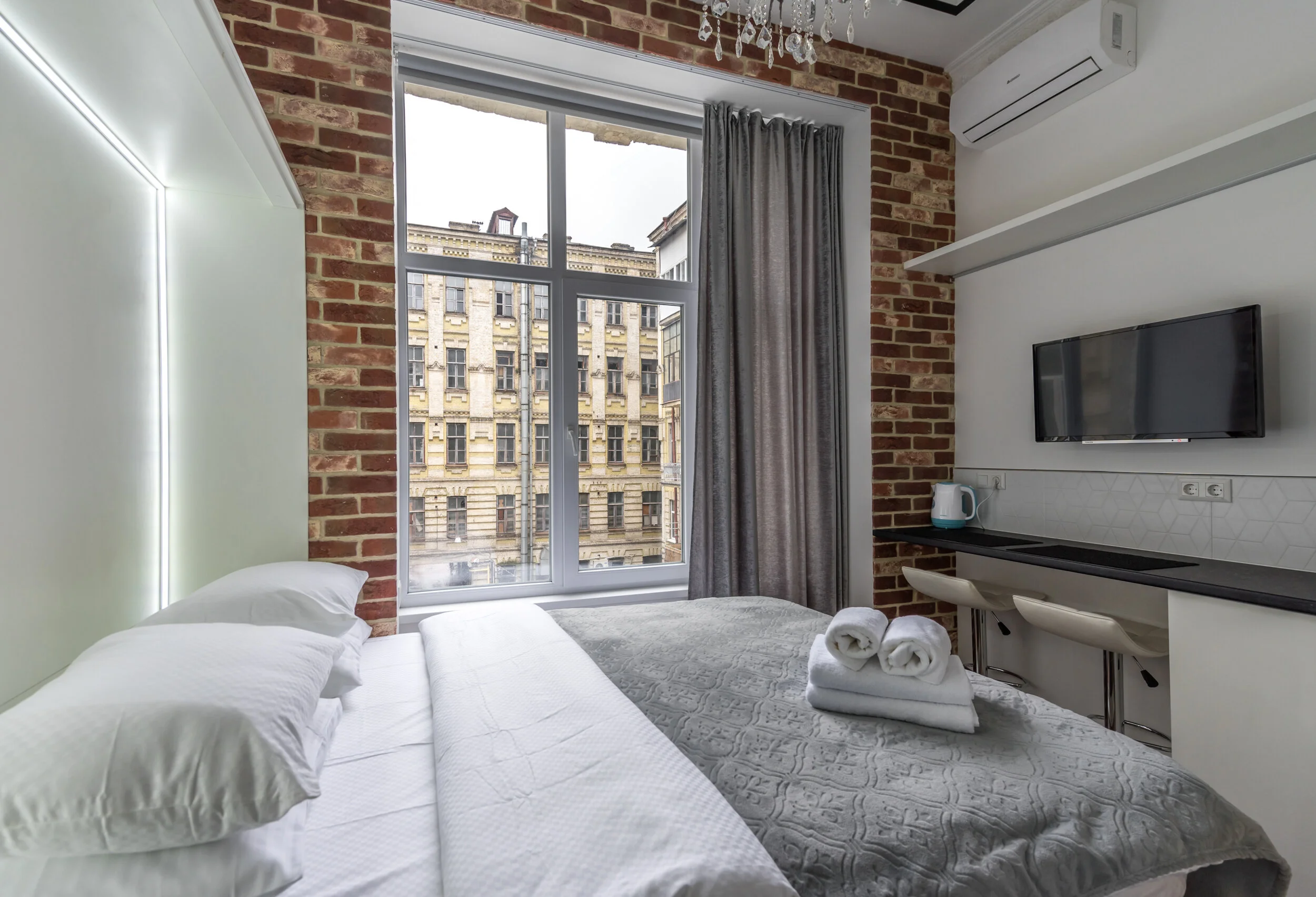 Modern hotel room with brick walls, a large window facing a historic building, a gray bedspread, stacked towels, a wall-mounted TV, and a small kitchenette with stools, kettle, and air conditioning unit.