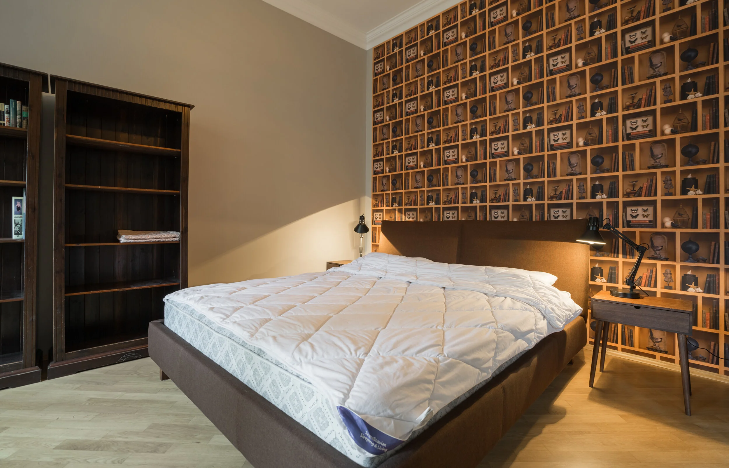 Modern bedroom with brown wood accents, a large bed with a white quilted comforter, empty dark wooden bookshelves, and wallpaper with a cube shelf design. A side table with a desk lamp is next to the bed.