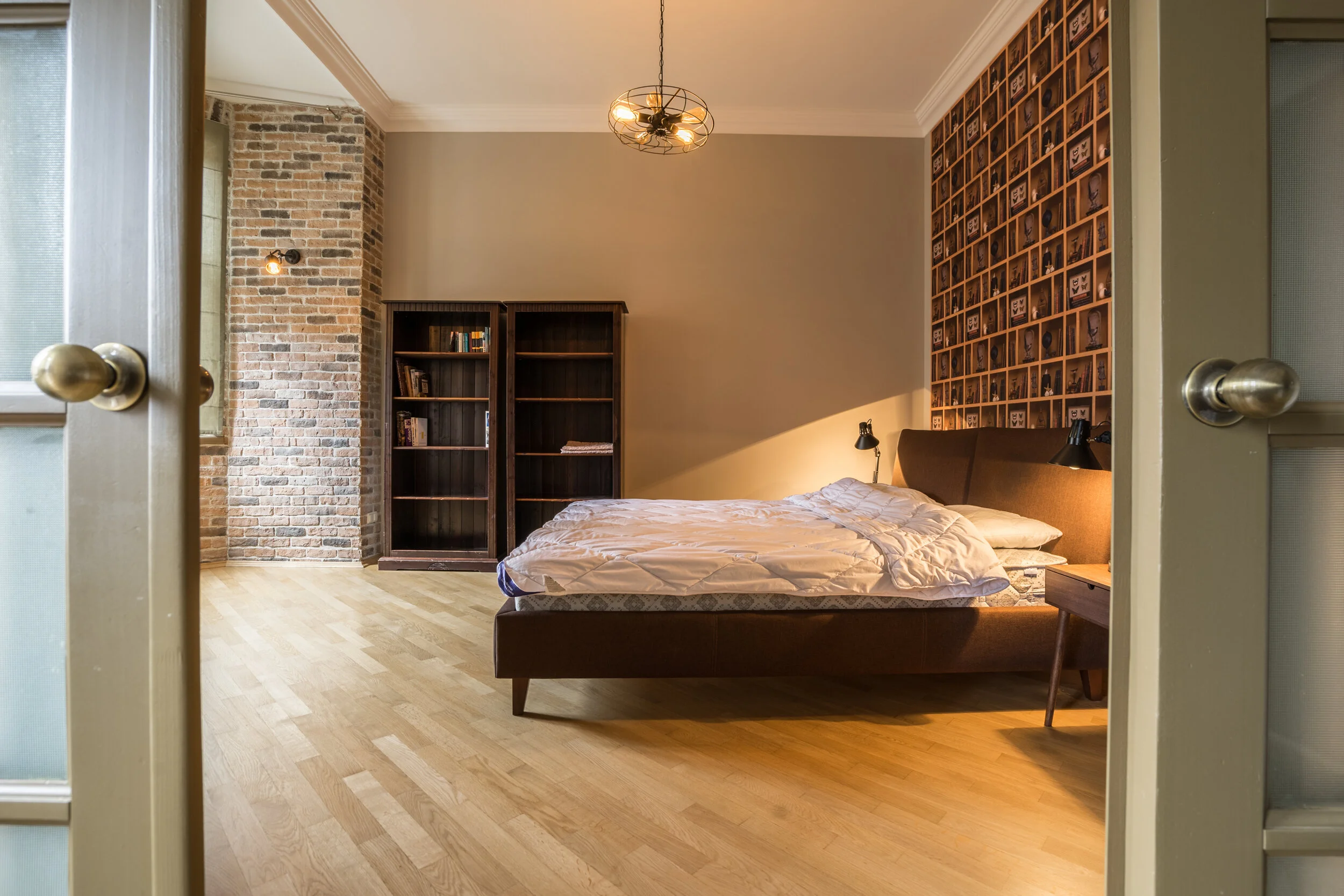 Cozy bedroom interior with a double bed, brick accent wall, wooden floor, open bookshelves, and a ceiling light fixture.