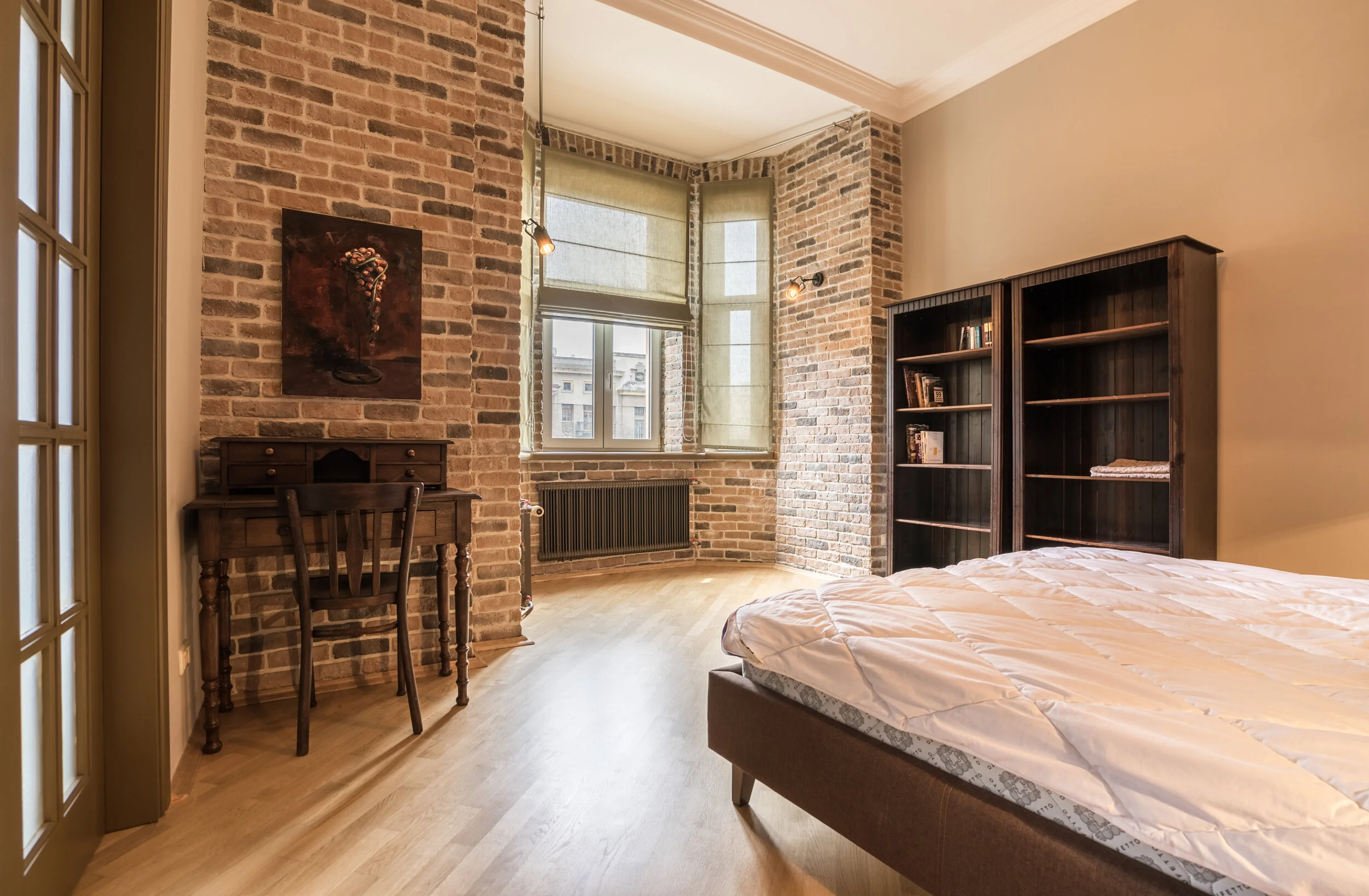 Bedroom with brick walls, wooden floor, a bed with white bedding, a wooden desk and chair, and a bookcase filled with books next to a large window with blinds.