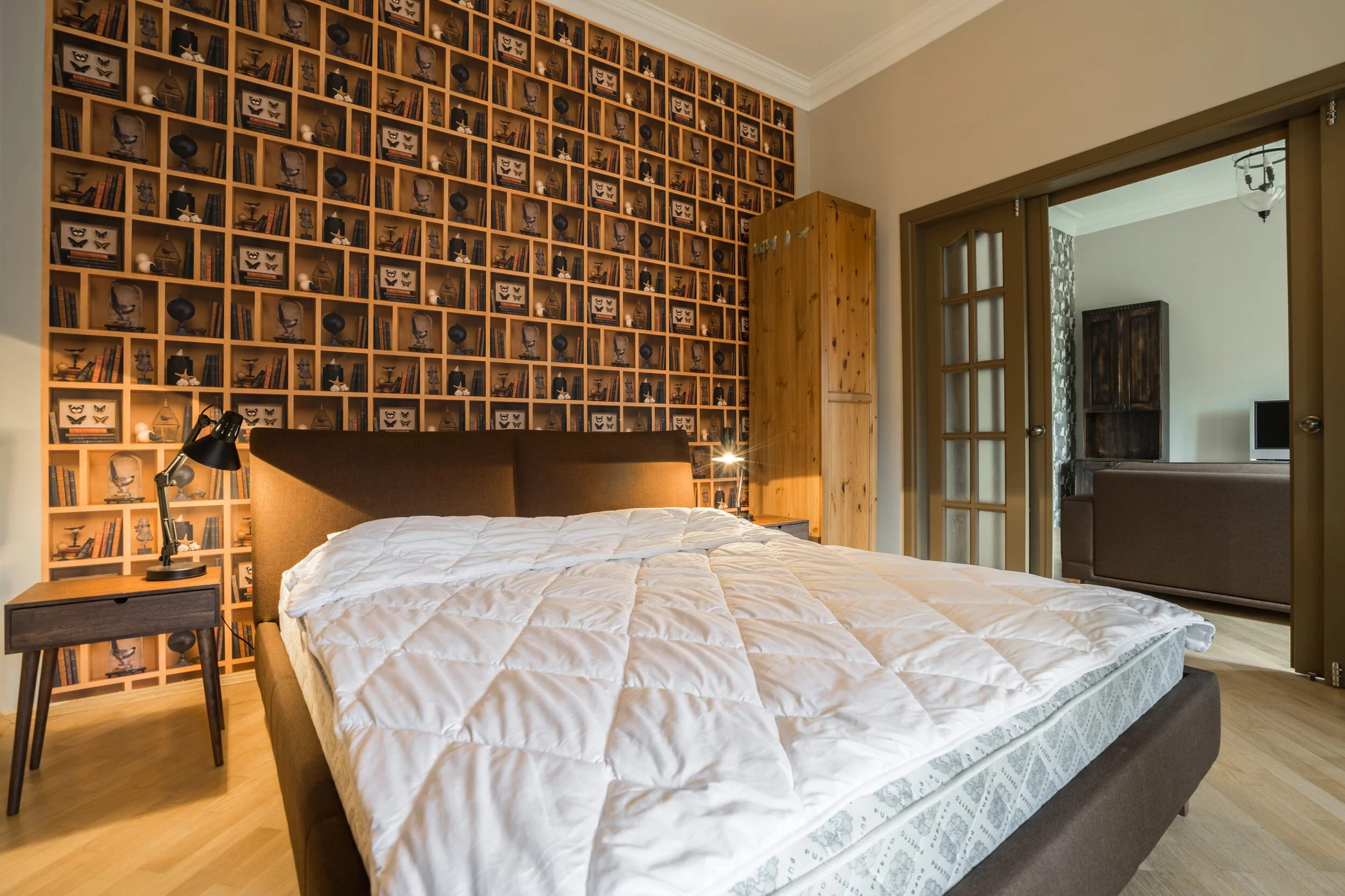 A bedroom with a quilted white comforter on a brown bed, a wooden nightstand with a black lamp, a unique bookshelf-patterned wallpaper, a wooden wardrobe, and sliding glass doors leading to another room with furniture.