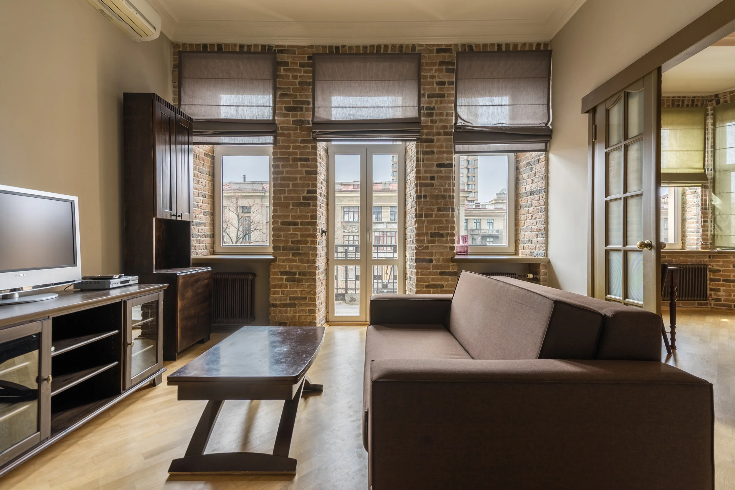 Living room with exposed brick walls, brown sofa, wooden coffee table, TV on a stand, and large windows with brown shades.