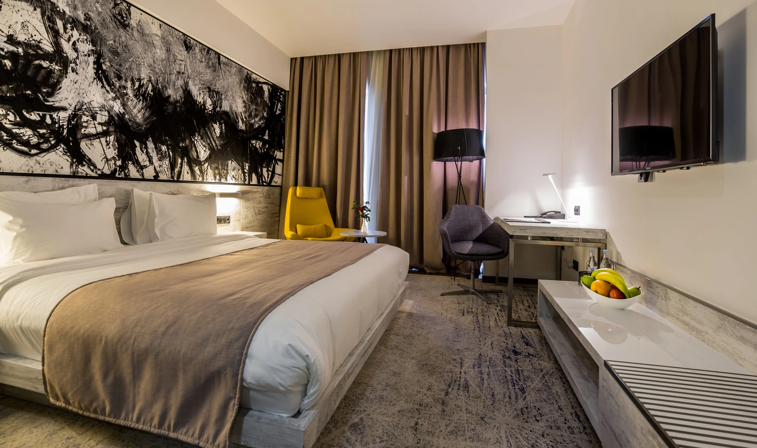 Modern hotel room with a large bed, abstract artwork, and cozy seating area with a yellow chair. The room has a desk, TV, and a fruit bowl on a side table.