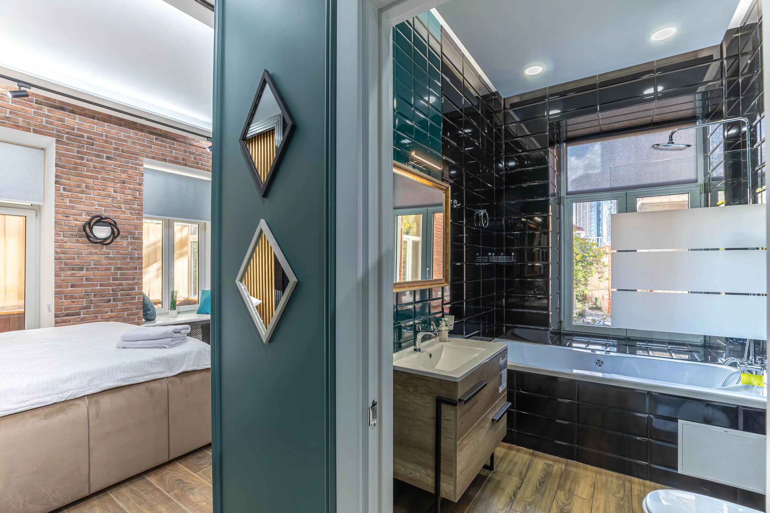 Modern apartment interior featuring a bathroom with black tile walls, a white bathtub, and a window, connected to a bedroom with brick walls and a neatly made bed.