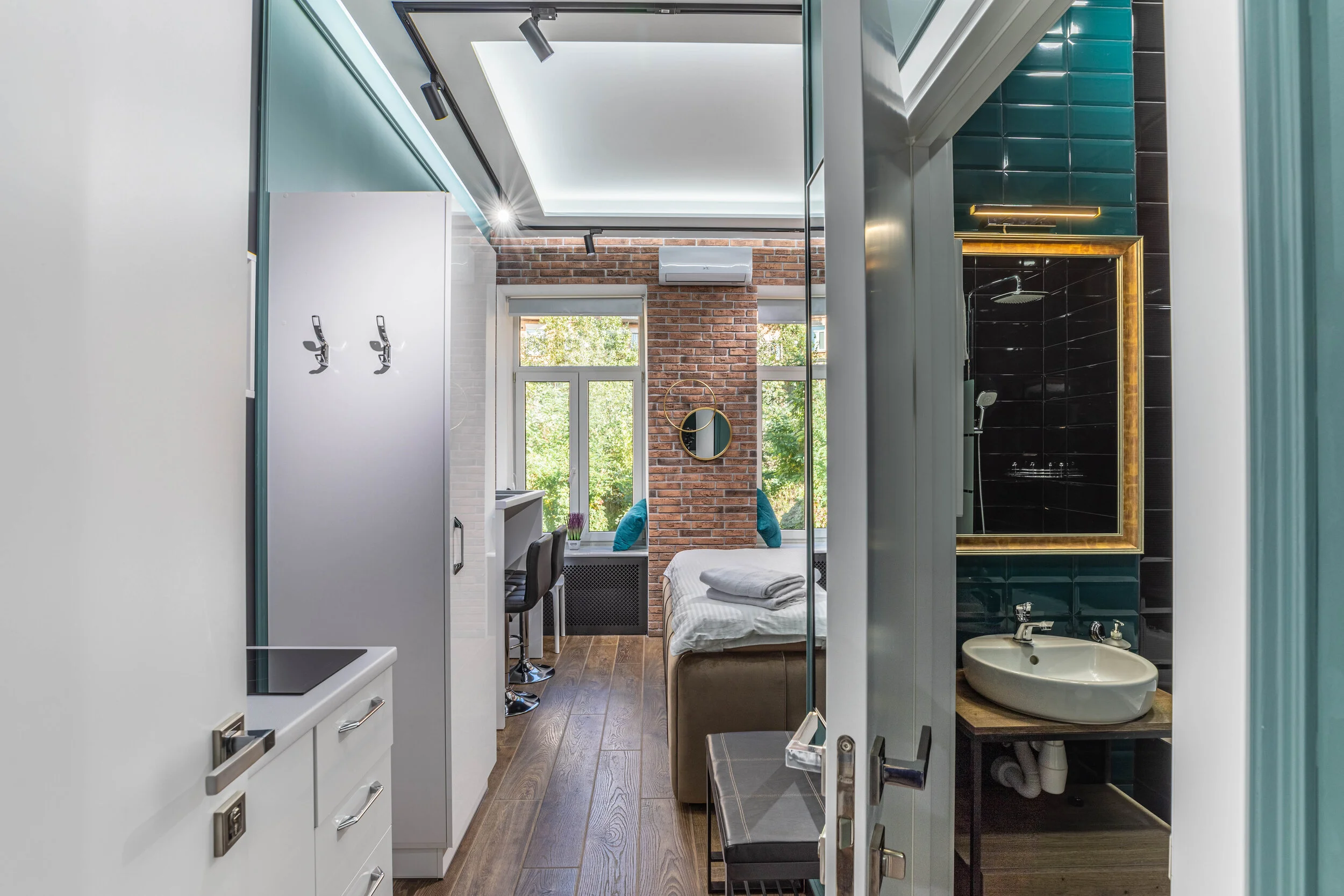 Modern apartment interior with open layout, featuring a small kitchen, living area, and bathroom. The kitchen has white cabinets and a stovetop. The living area includes a couch, a table, and brick accent wall with large windows. A bathroom with a si