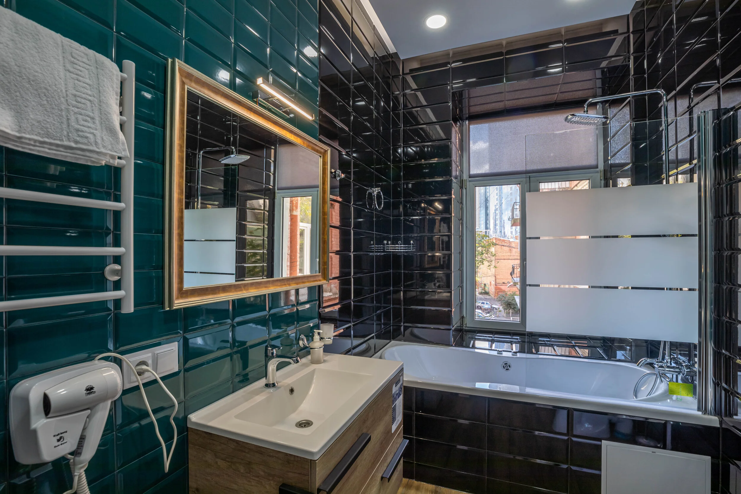 Modern bathroom with dark tiles, a large mirror, white sink, bathtub, and exterior window.