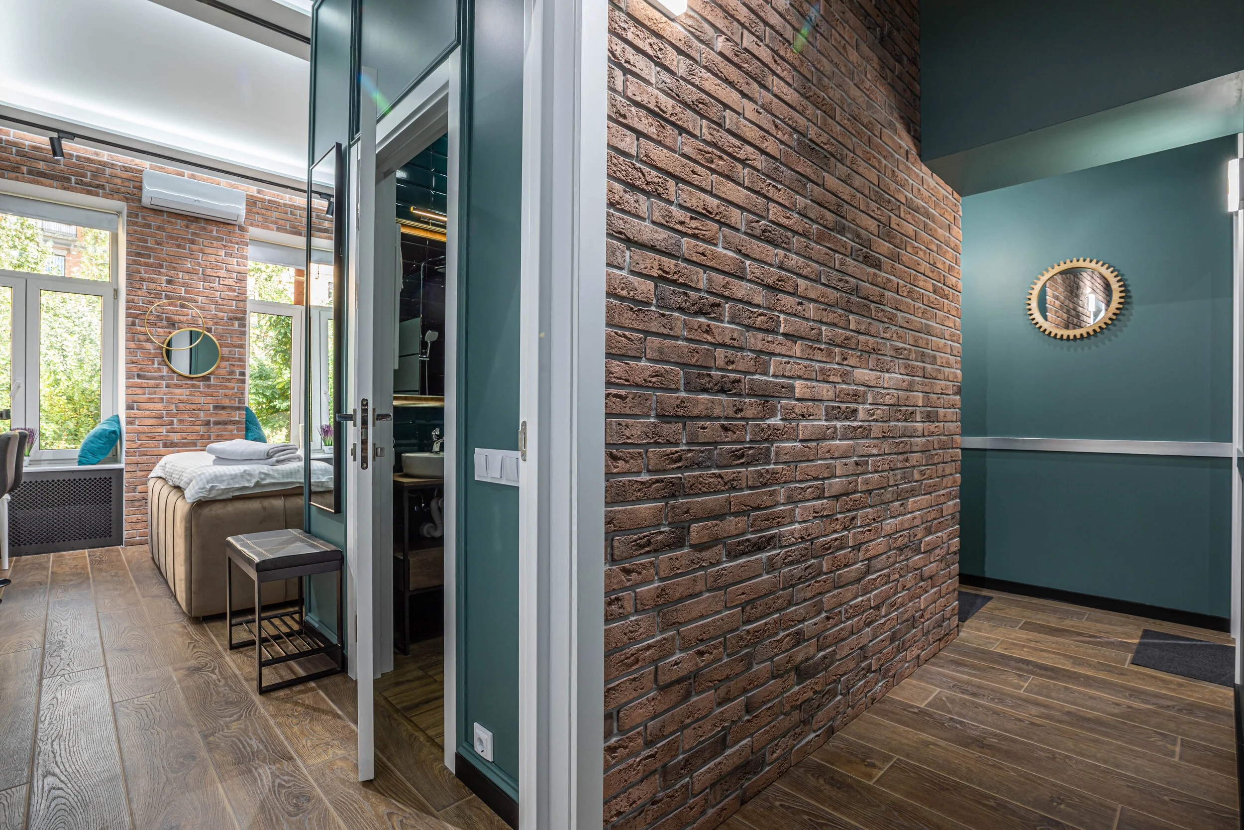 Modern apartment interior with exposed brick walls, wooden flooring, and teal-painted sections. The room includes a bed with cushions, a mirror, and natural light from large windows.