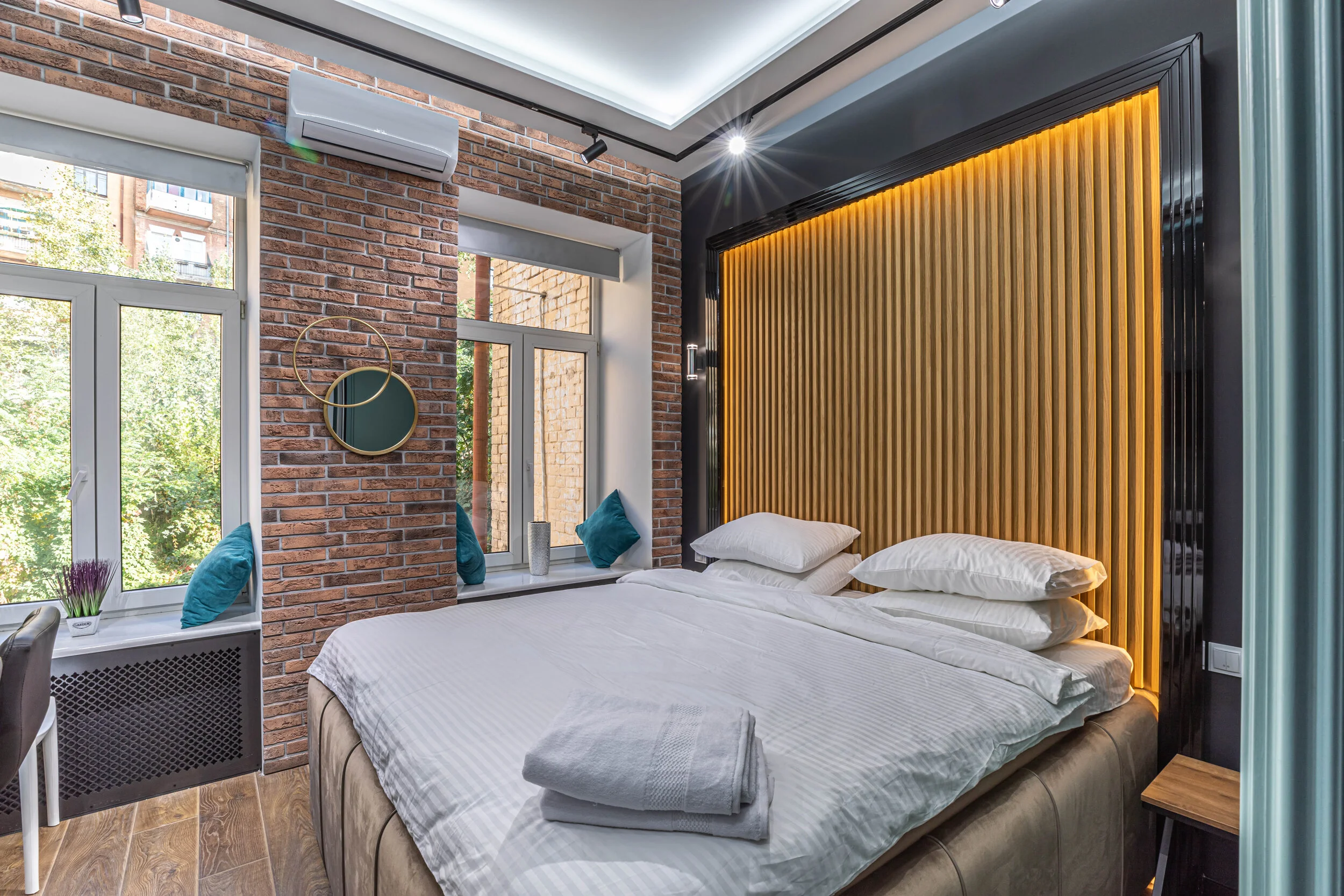 Modern bedroom interior with a double bed, white bedding, striped pillows, a wooden slatted feature wall with warm lighting, brick walls, large windows with teal cushions on the sill, and overlapping circular mirrors. An air conditioning unit is moun