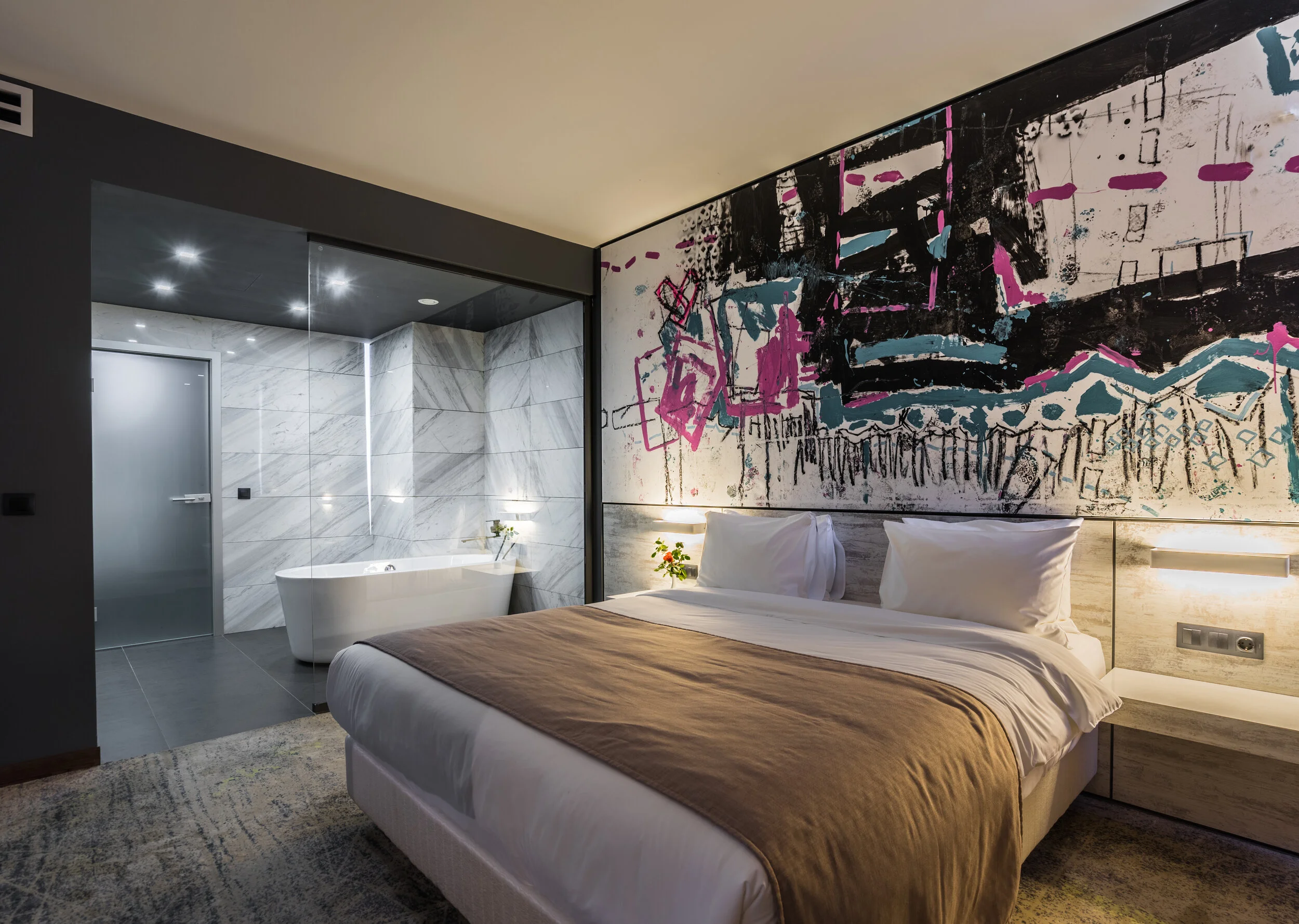 Modern hotel room with a large bed, abstract wall art, glass-enclosed bathroom featuring a marble tub and walls, and soft ambient lighting.