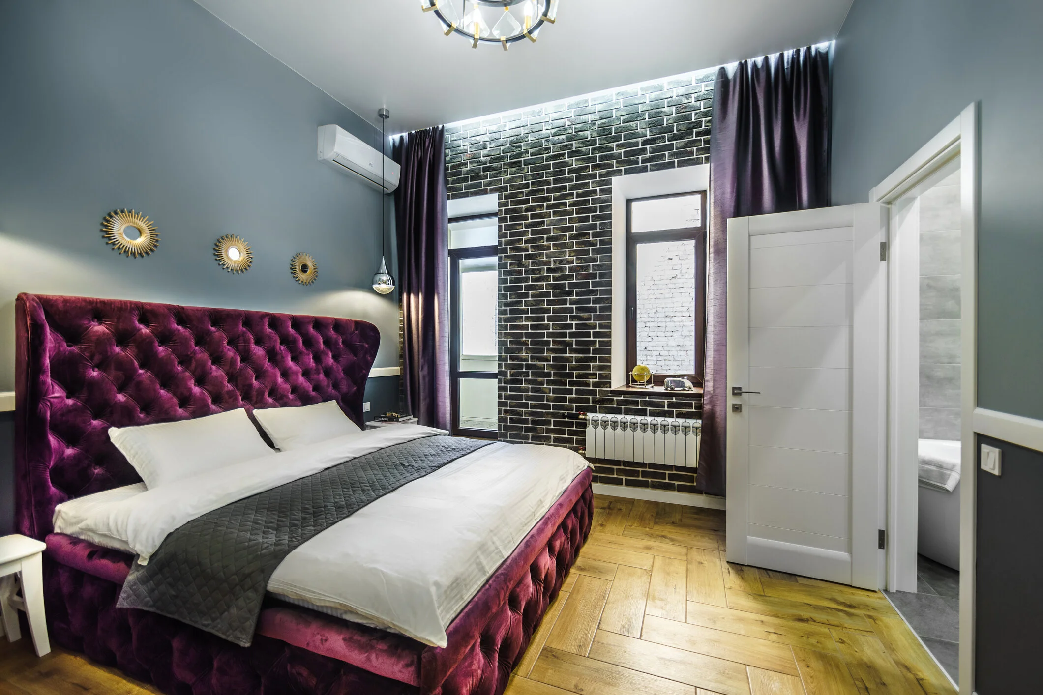 Modern bedroom with a purple upholstered bed, brick accent wall, parquet flooring, and decorative wall mirrors.