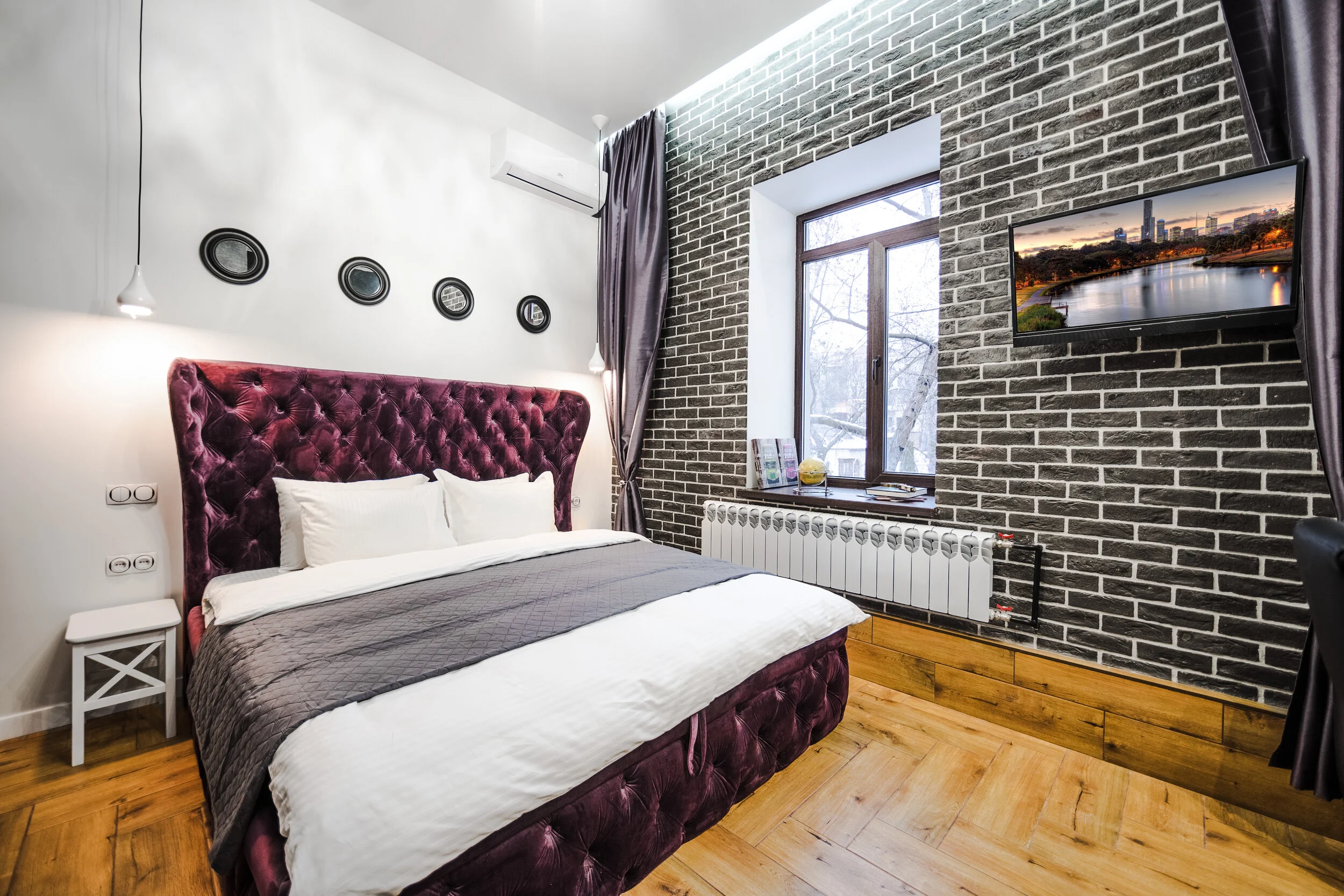 Modern bedroom interior with gray brick wall and large window. Features a purple velvet headboard, white bedding, wall-mounted TV, decorative wall plates, and wooden flooring.