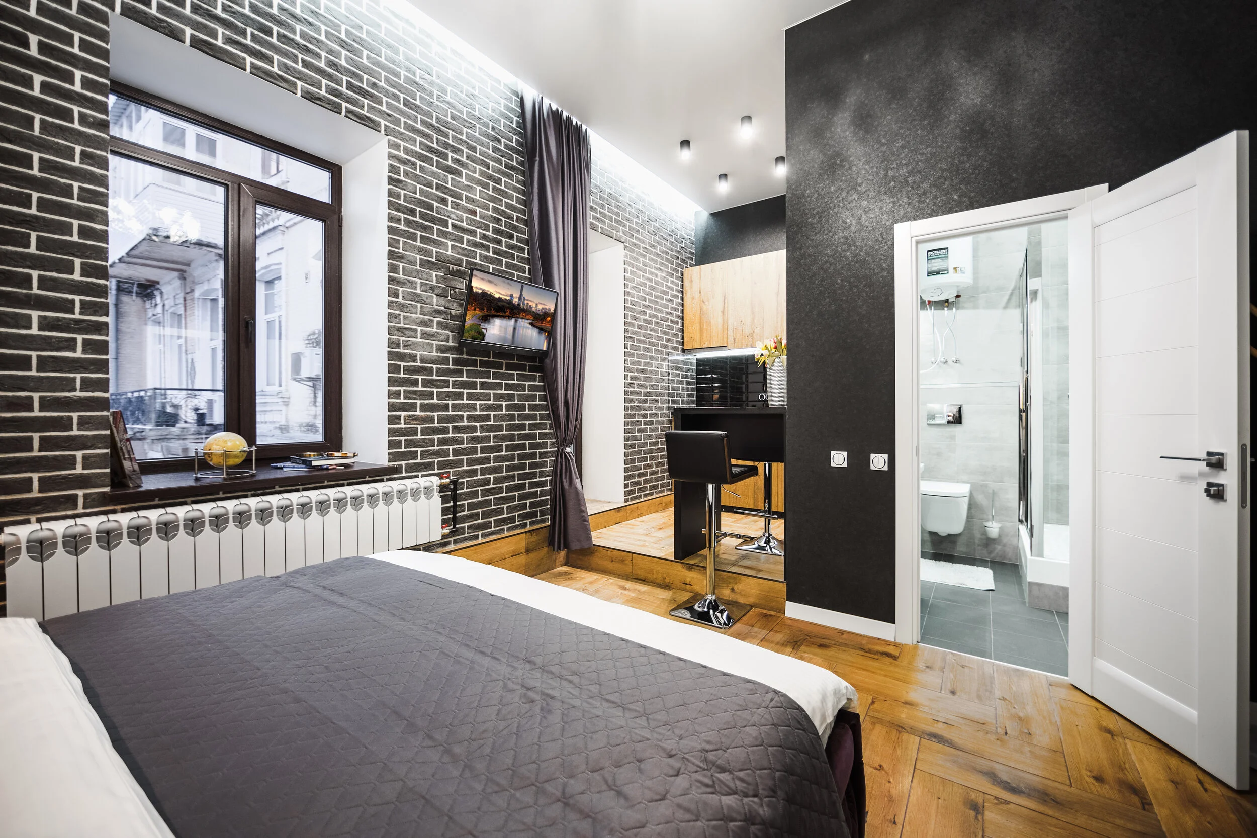 Modern bedroom with brick accent wall, large window, bed with gray comforter, flat-screen TV, wooden floor, bar stool, small kitchen area, and en suite bathroom with open door.