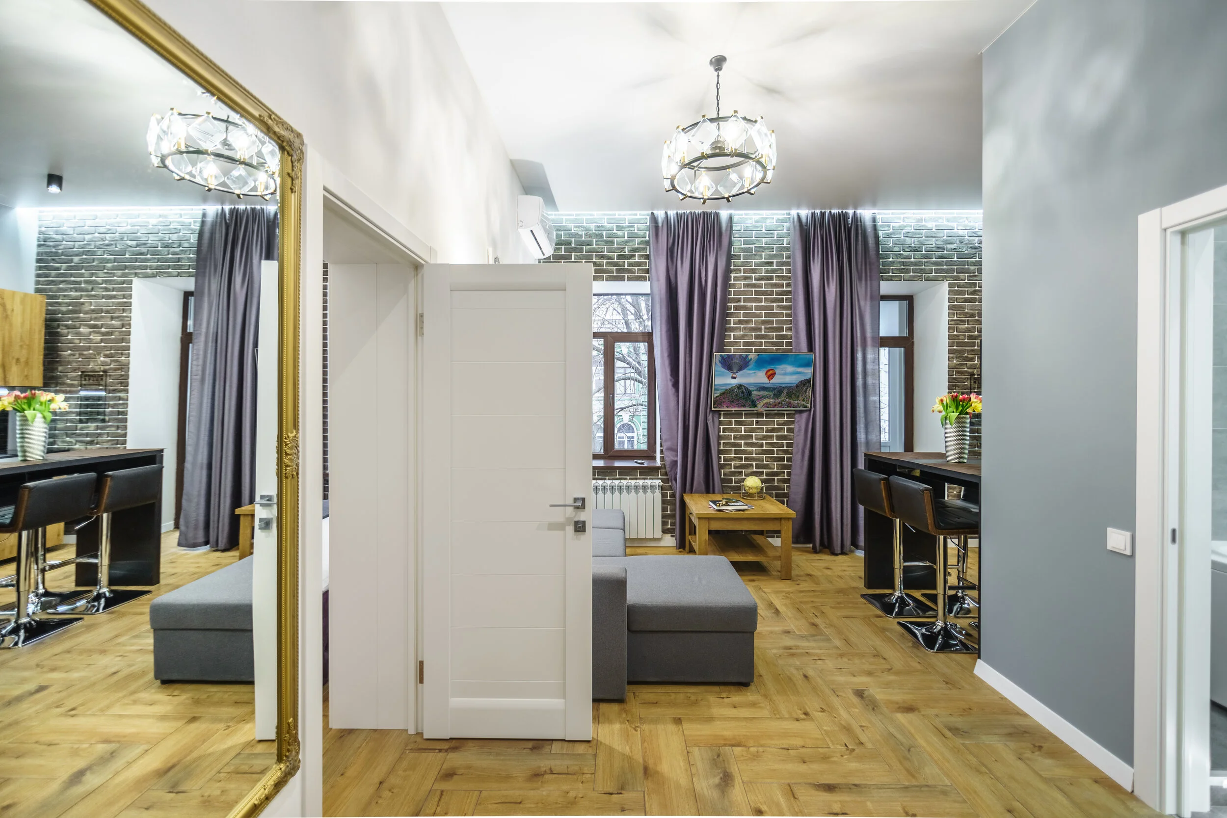 Modern living room interior with gray and brick walls, wooden flooring, a gray sofa, a coffee table, tall bar stools, floral arrangements, and a large mirror.