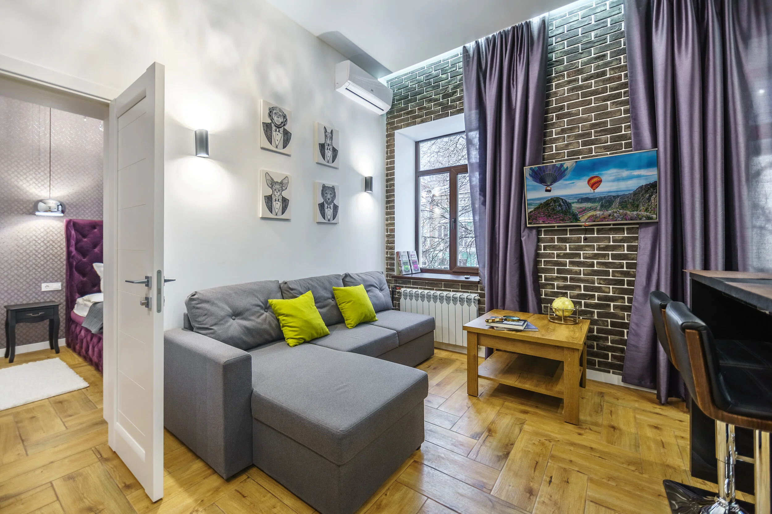 Modern living room with gray sofa, green pillows, wall art of animals in suits, wooden coffee table, and a picture window with long curtains. View of bedroom through open door.