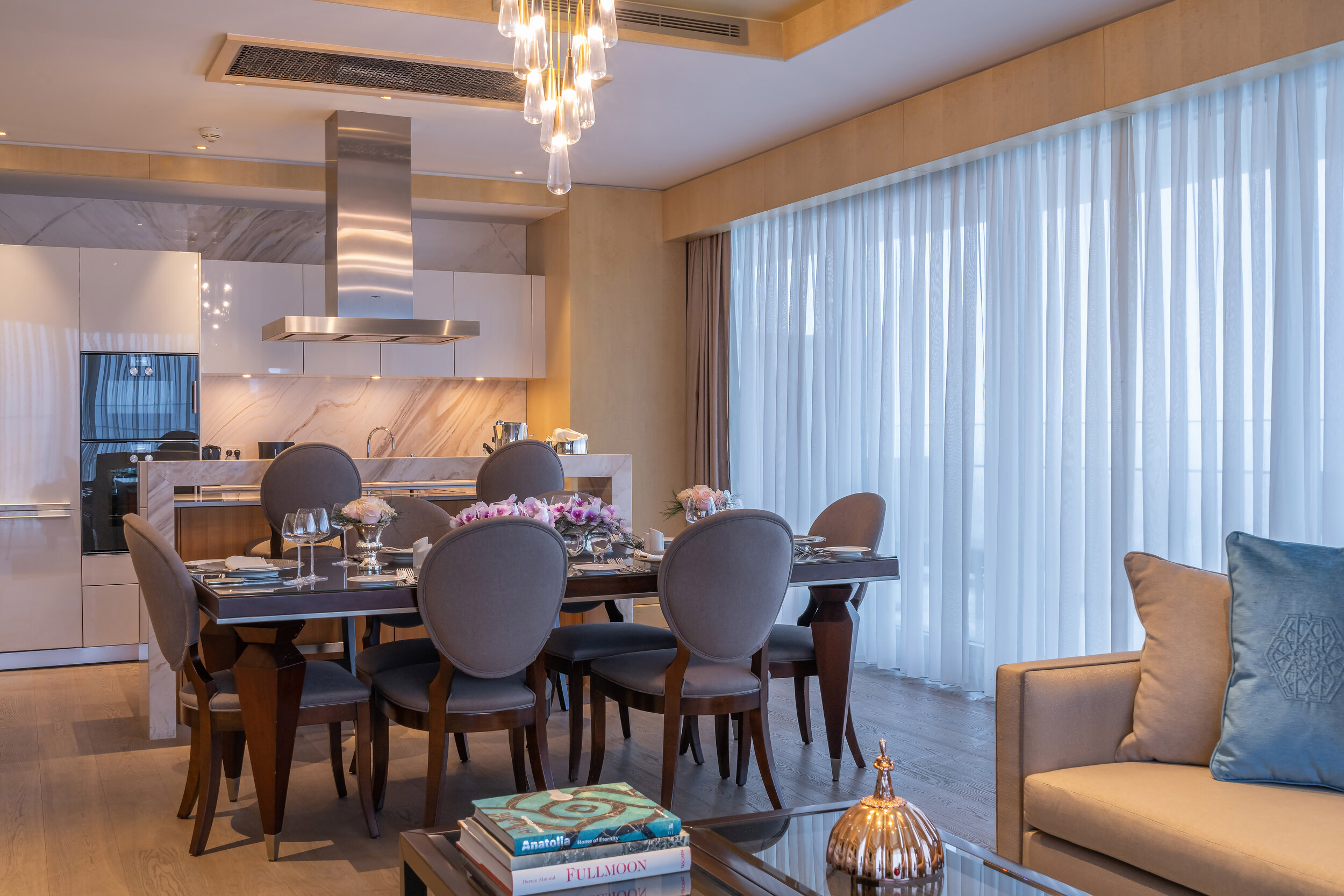 Modern dining area and kitchen with a table set for six, elegant chairs, a chandelier, and large windows with sheer curtains.