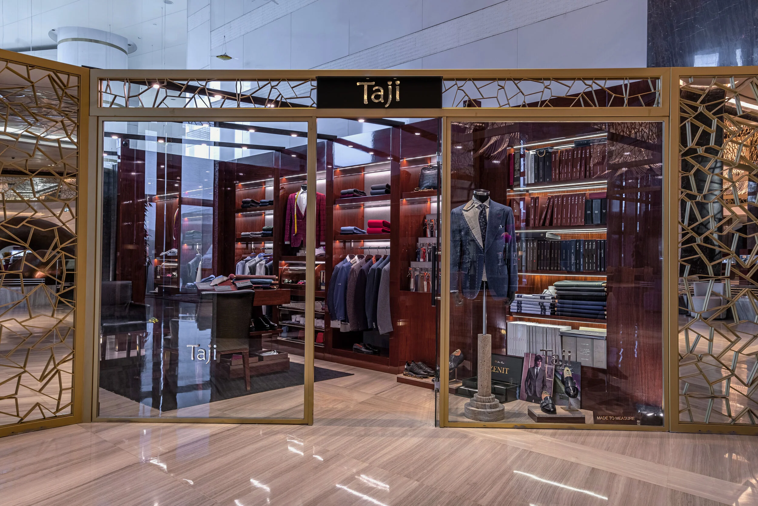 A luxury clothing store named 'Taji' with suits and accessories displayed in a glass-fronted shop, featuring elegant wooden shelving and mannequins dressed in suits.