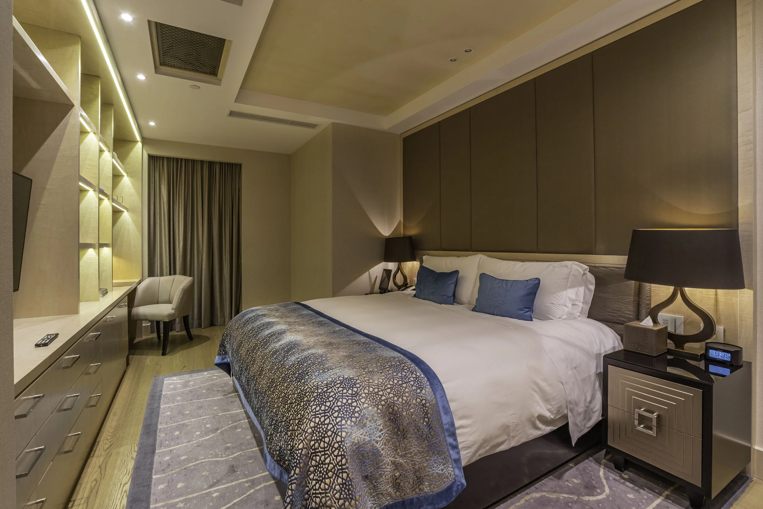 Modern hotel room interior with a large bed, blue cushions, elegant bedside tables, decorative lamps, built-in shelving, and a chair by closed drapes.