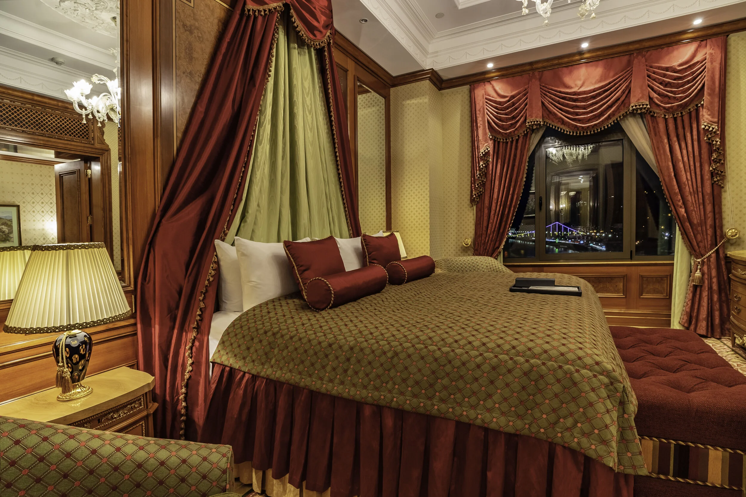 Luxurious hotel room with an elegant, canopy-style bed featuring red and green drapes and pillows. A large window with red curtains offers a view of a brightly lit bridge. The room has wood-paneled walls, ornate light fixtures, and a bedside lamp.