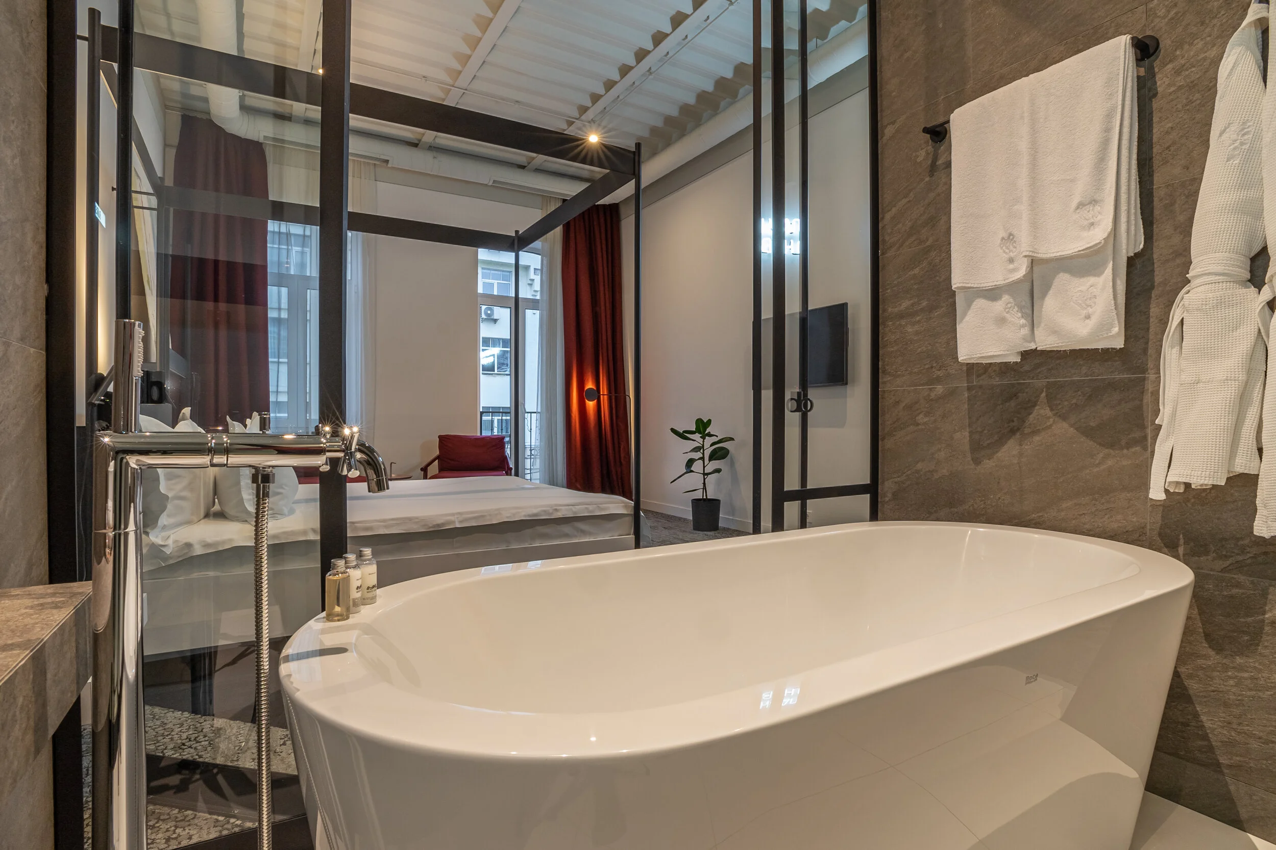 Modern hotel room with freestanding bathtub, glass partition, and view of bedroom with a four-poster bed, red armchair, and a potted plant.