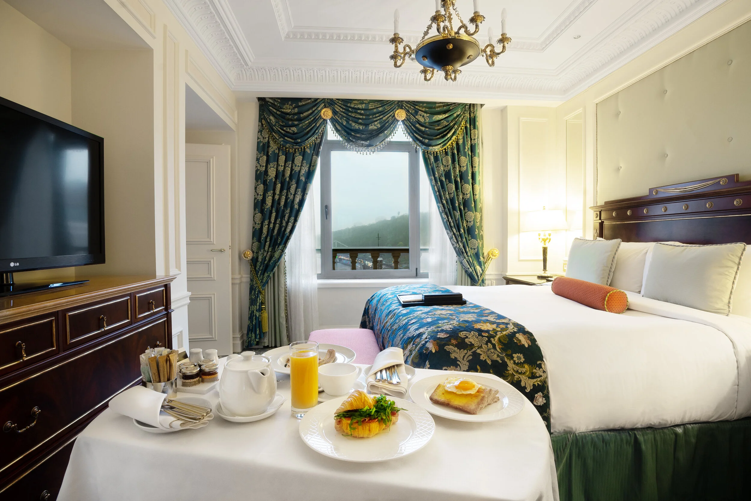 Elegant hotel room with a king-sized bed, ornate curtains, and a chandelier. A breakfast tray on a table features orange juice, a teapot, toast with an egg, and a croissant with salad. A flat-screen TV sits on a wooden dresser.