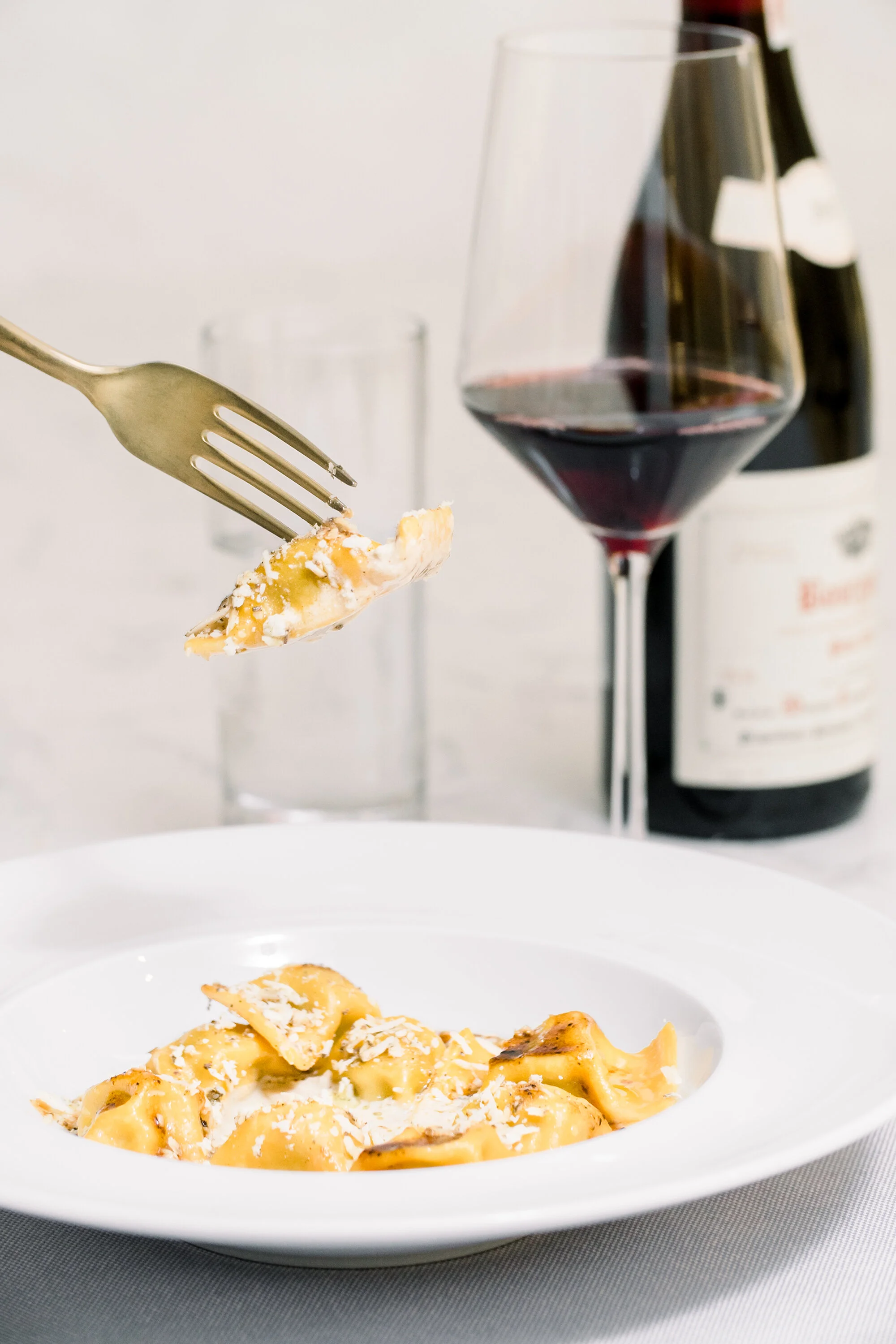 Plate of ravioli with grated cheese and a fork, next to a glass of red wine and a wine bottle.