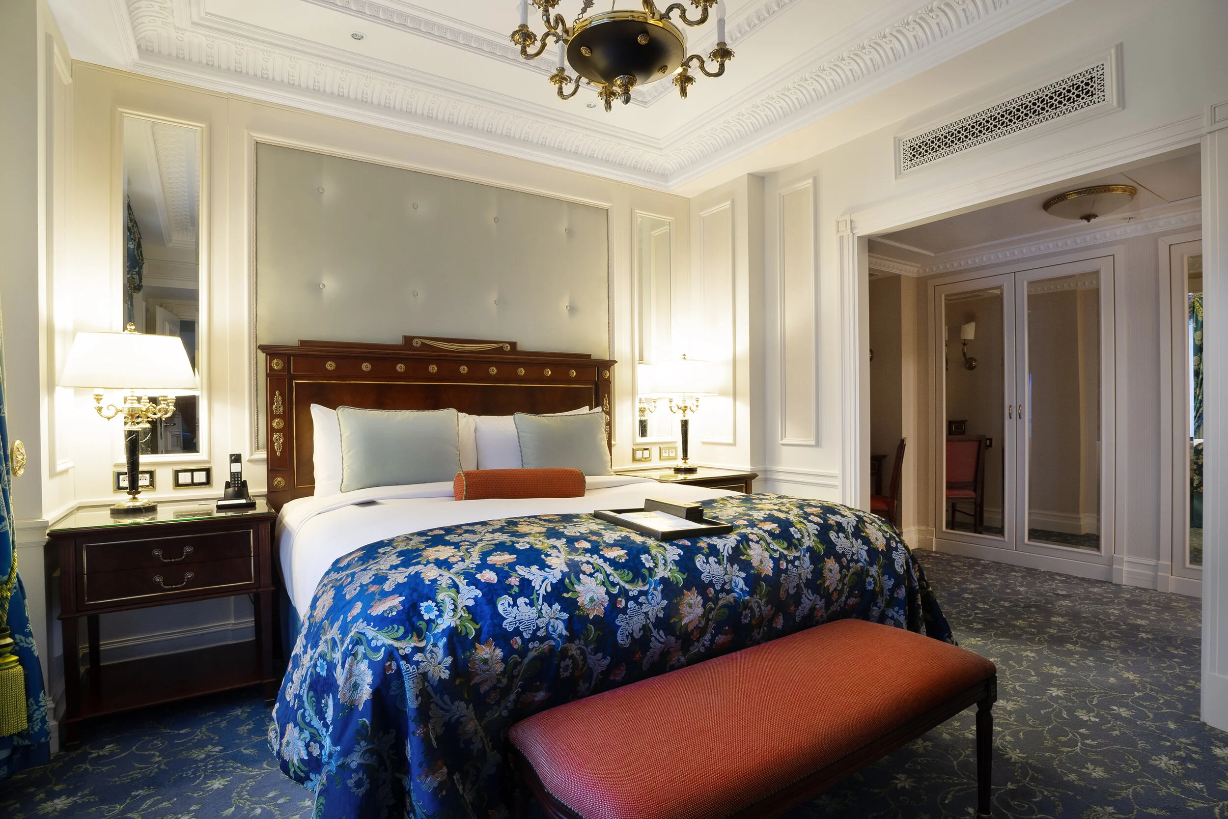 Luxury hotel room with a king-sized bed, ornamental headboard, floral bedspread, nightstands, lamps, mirrored closet doors, and elegant decor.