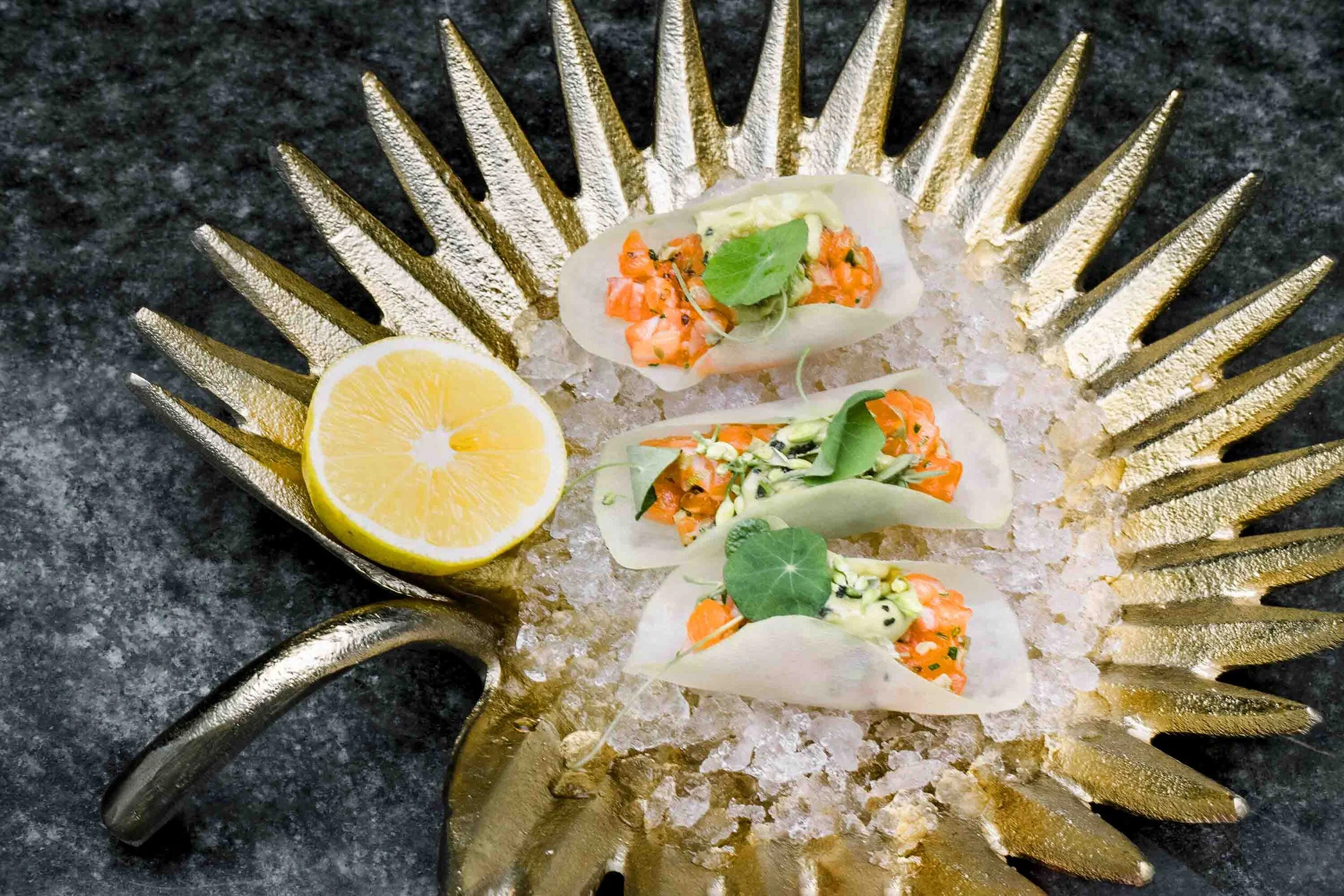 Three pieces of sushi with salmon and green garnishes on a decorative golden plate with crushed ice and a lemon half.