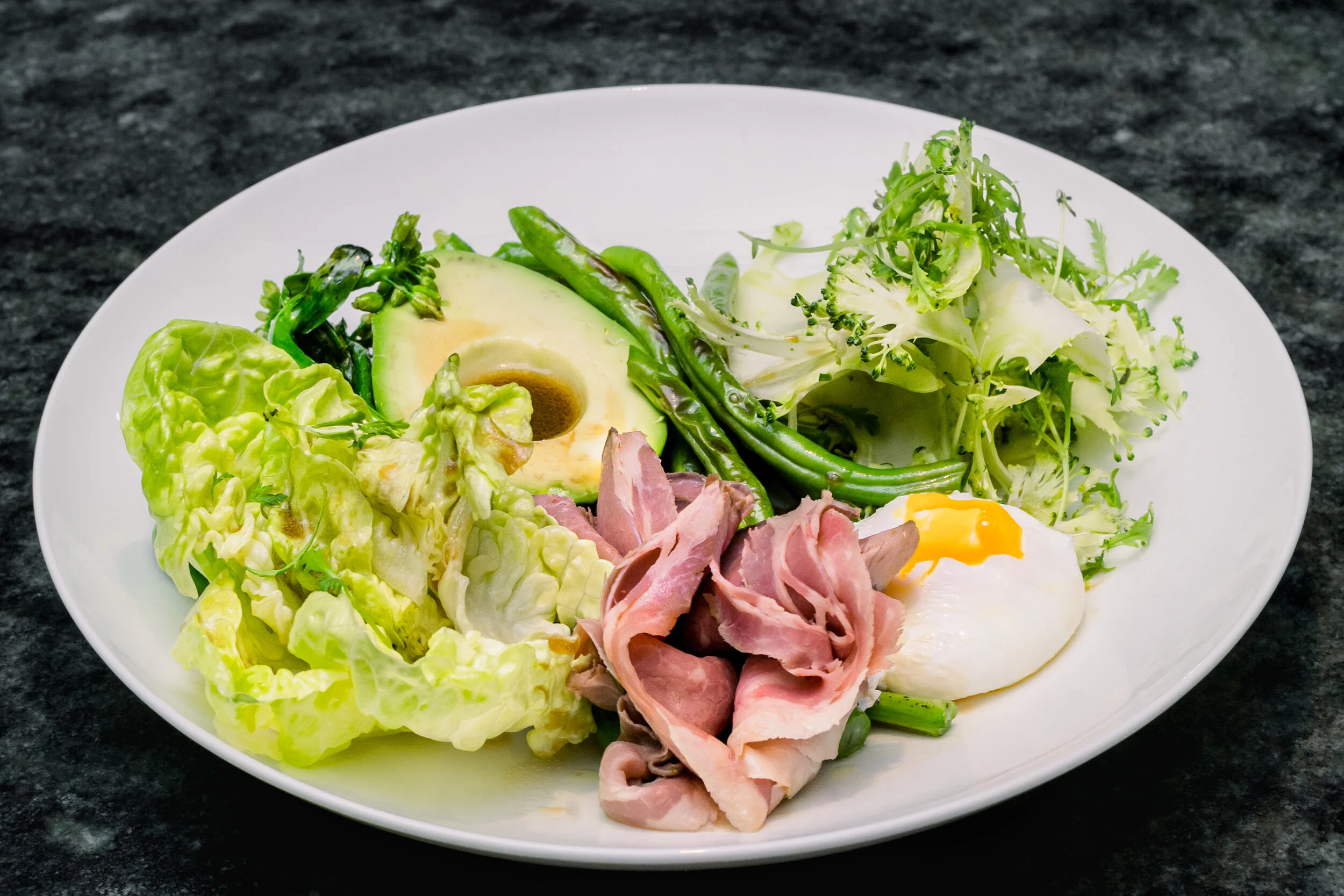 Plate with salad including lettuce, avocado, green beans, poached egg, and ham slices on a dark background.