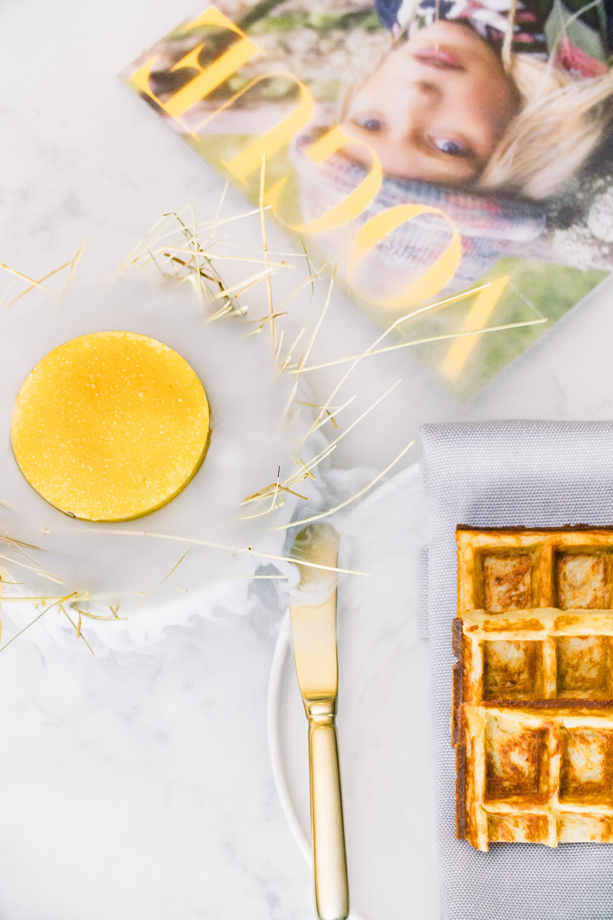 A round yellow pastry on a marble surface with a magazine, a gold knife, and half a square waffle on a gray napkin.