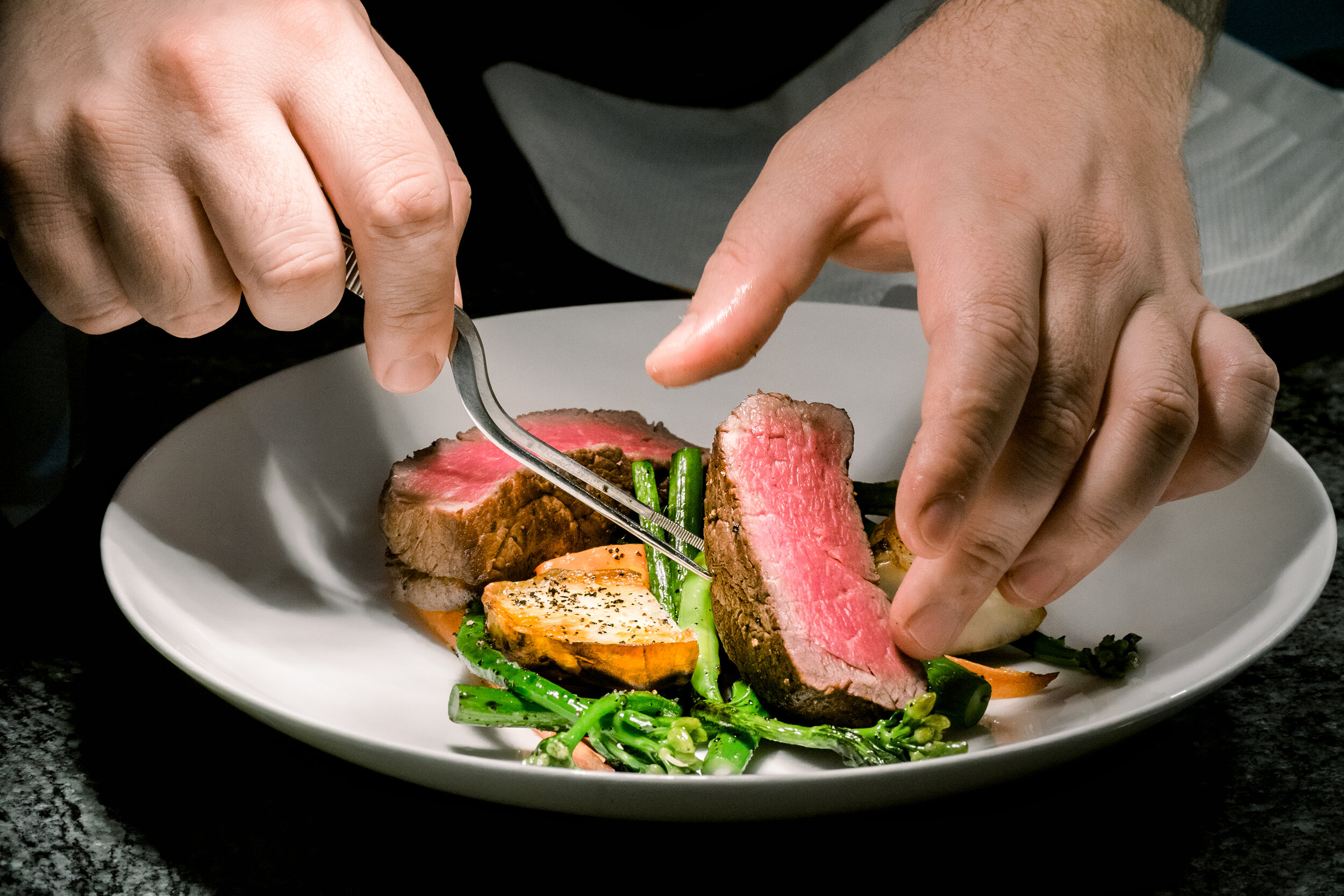 Chef plating medium-rare beef steak with grilled vegetables on a white plate.