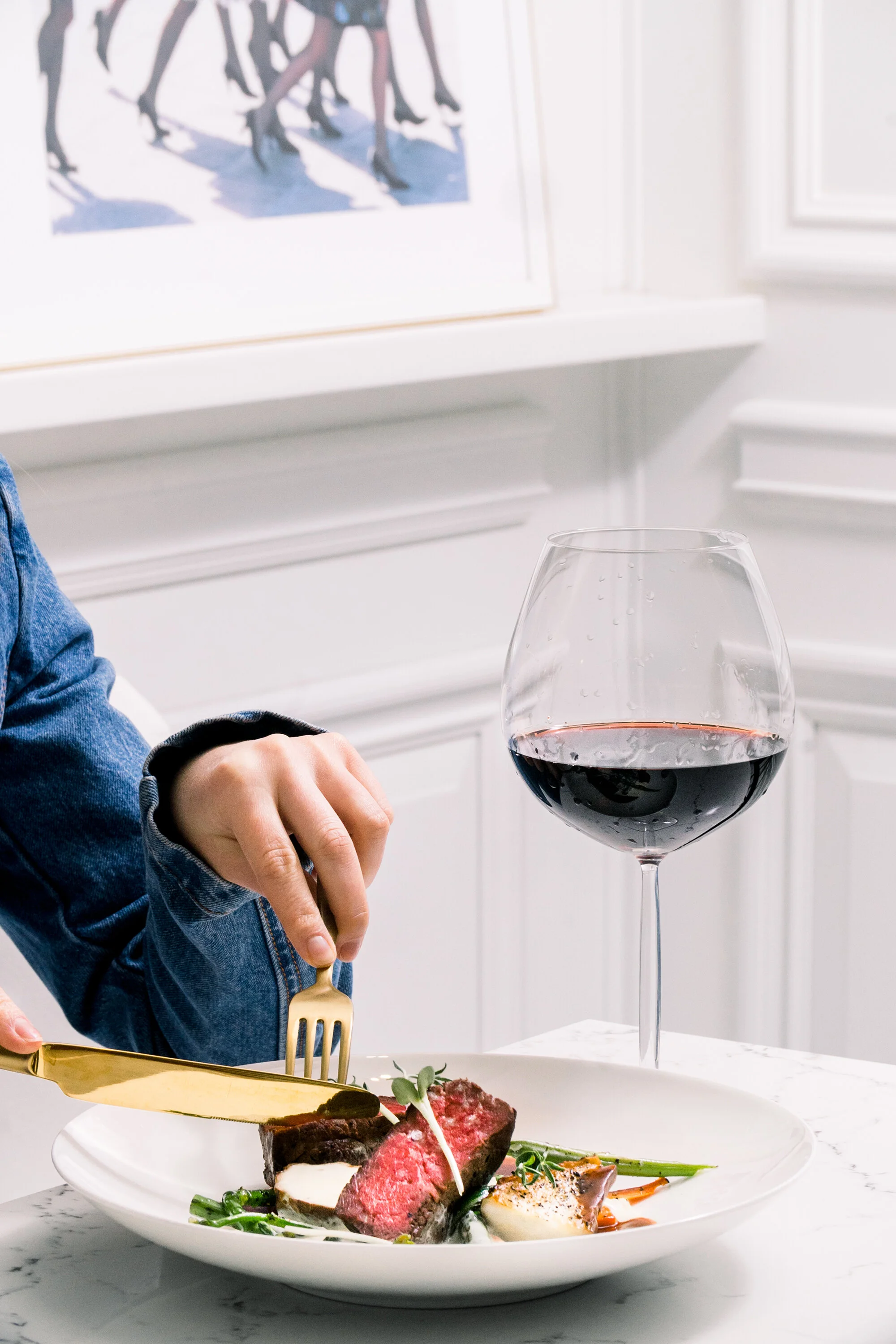 A person cuts a piece of steak on a plate with vegetables, next to a glass of red wine, in a white setting.